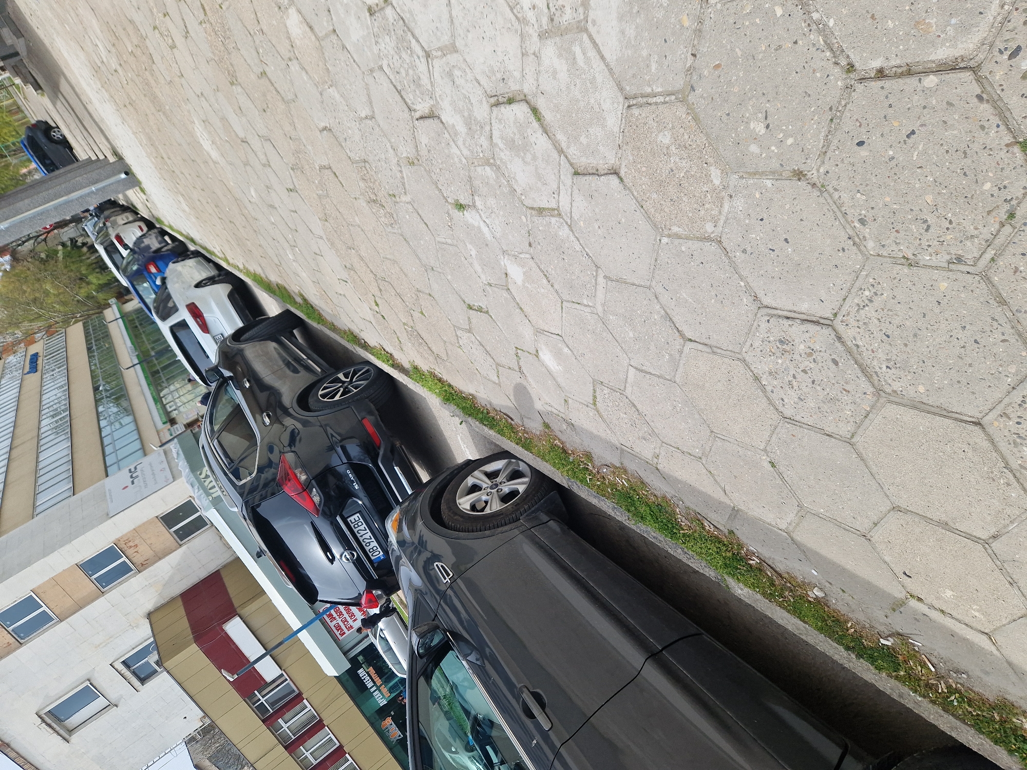 The image depicts a street scene in Lovech, Bulgaria, with several cars parked along the roadside. The cars are of various colors and models. A building with a sign that reads "JOYS," "SUL," and "ДРЕХИ И МЕБЕЛИ" (Clothes and Furniture) is visible. The license plate "OB 9212 BE" is on one of the cars. The environment consists of buildings, sidewalks, and a concrete retaining wall.