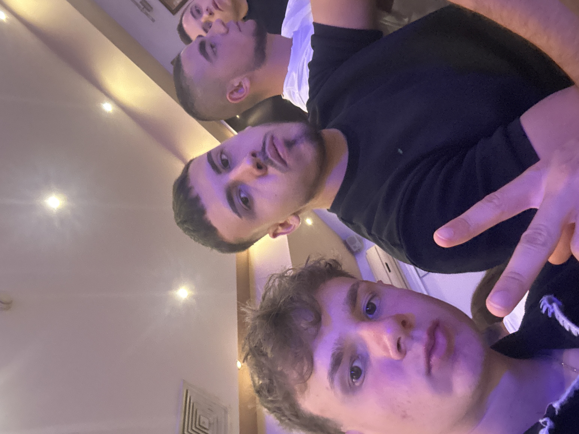 A selfie, taken at an upward angle, captures three young men indoors. The foreground figure has light skin, short brown hair, and is wearing a light-colored shirt. The middle figure has olive skin, short dark hair, a trimmed beard, and is wearing a black shirt. The background figure has olive skin, short dark hair, and is wearing a white shirt. The middle figure is making a duck-face expression. The foreground figure is making a peace sign with his fingers. The location is indoors, possibly a home or establishment in an unknown city. No specific evidence exists to establish that the photo was taken in the city of Elhovo, Bulgaria. The lighting is diffused and the background is partially obscured. A ceiling vent can be seen in the bottom left corner of the image.
