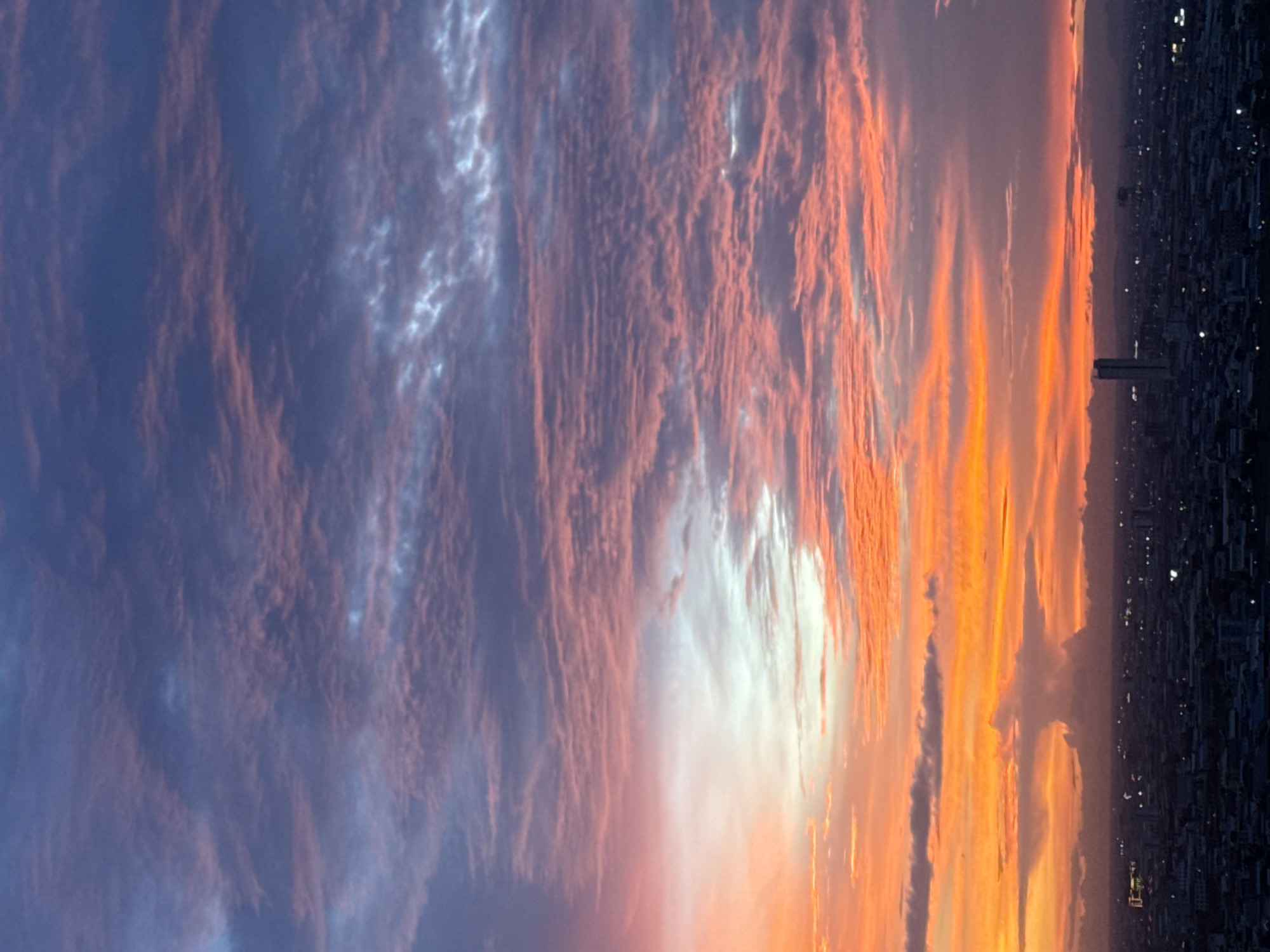 The image depicts a dramatic sunset over the cityscape of Khan Mean Chey, Cambodia. The sky is filled with a vibrant palette of colors, transitioning from fiery oranges and yellows near the horizon to soft pinks and purples, and finally to deep blues at the zenith. Wispy clouds are scattered across the sky, catching the light and creating a textured, almost painted effect.
Below the striking sky, the urban landscape of Khan Mean Chey is visible as a dark silhouette against the colorful backdrop. Buildings, including at least one tall, prominent skyscraper, are discernible as indistinct shapes. The lights of the city twinkle, suggesting it is either dusk or the early evening. A notable feature on the horizon is a large, cloud formation that resembles a mushroom or an anvil, a possible sign of distant weather activity. The overall scene is one of natural beauty juxtaposed with the man-made environment of the city.