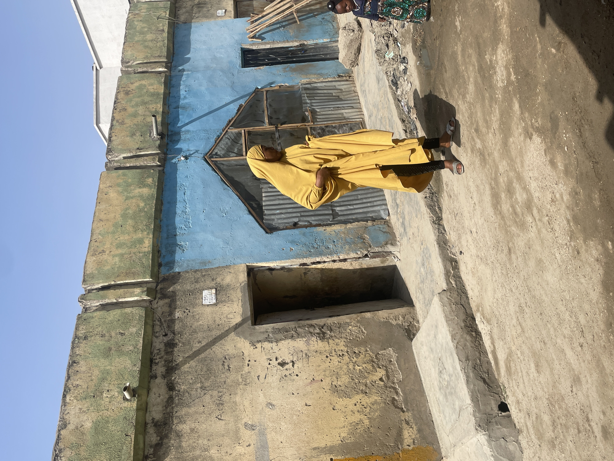 A woman in a full-length yellow garment is standing in front of a weathered, multi-colored building in Unknown, Unknown. The building's facade is a mix of green, blue, and beige tones, appearing aged and somewhat deteriorated. There are multiple openings or windows in the building, some with metal coverings. The woman is facing forward and has a hooded head covering that is part of the yellow garment. The ground is sandy and uneven. Part of another person is visible in the upper right corner, wearing a dark patterned dress. The sky is visible in the upper left and is a light blue color.