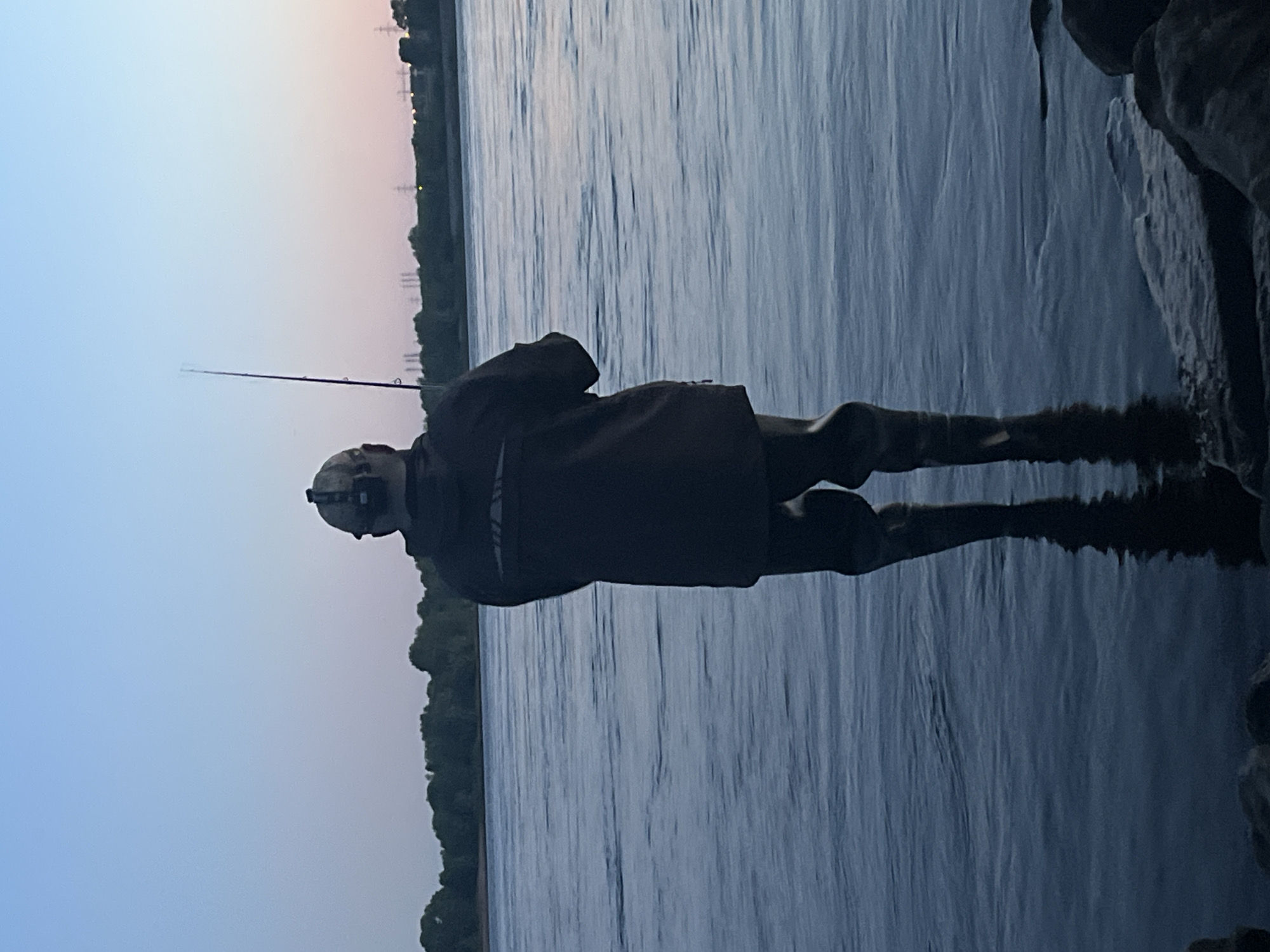 The image shows a person fishing in a body of water, likely the Kama River, at dusk. The person is wearing waders and a jacket and is holding a fishing rod. The water is calm, and the sky is a pale blue with hints of pink. The background shows a treeline and some industrial structures. The scene appears to be in the city of Chaikovsky, Russia, given the context.