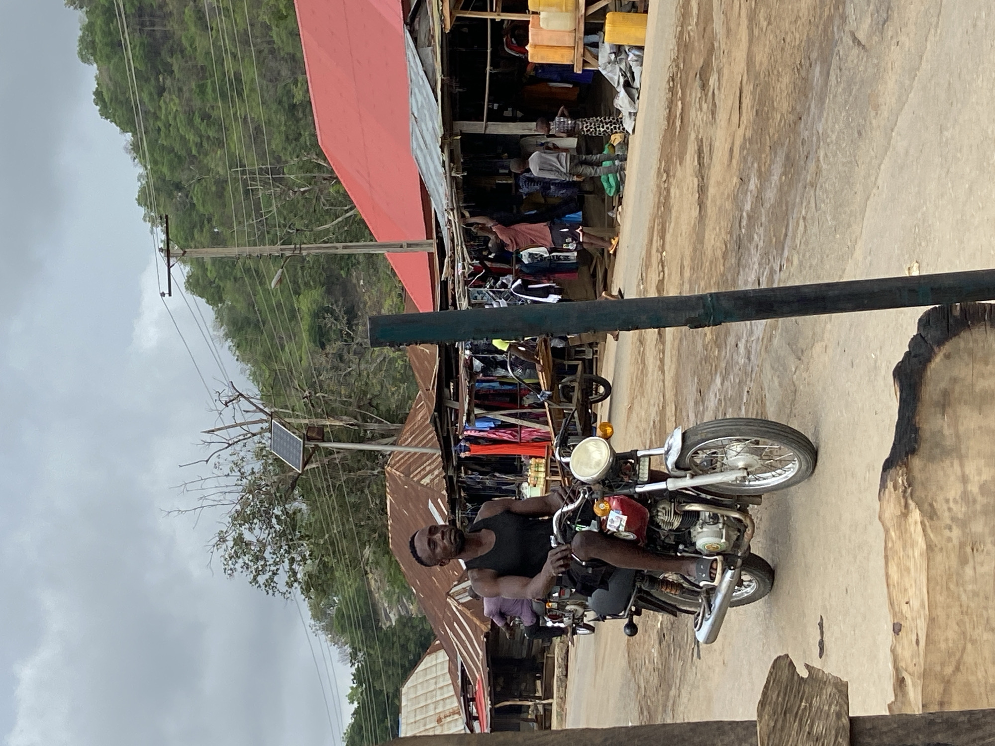 This image, set in Mararaba, Nigeria, captures a vibrant street scene during the daytime under an overcast sky. In the foreground, a man with dark skin, wearing a black tank top, is seated on a red and silver motorcycle. He looks directly towards the viewer with a neutral expression, his hands resting on the handlebars. The motorcycle features a prominent round headlight and visible engine components.
Behind the man, the scene transitions to a bustling market area. Several people are visible near open-fronted market stalls, which display an array of goods, including hanging clothes and items stored in stacked yellow and white jerry cans. The market structures are characterized by buildings with corrugated metal roofs, some painted red and others showing signs of rust.
In the mid-ground, utility poles support a network of power lines. A notable detail is a small solar panel mounted on one of these poles, suggesting a localized power solution. The background is dominated by lush, green, forested hills or mountains, indicative of a natural landscape bordering the settlement. The ground beneath is a mix of dirt and uneven pavement. Wooden planks are visible in the immediate foreground, potentially part of a nearby structure or barrier. The overall scene depicts a typical day in a busy, semi-urban market environment. No discernible text is visible in the image.