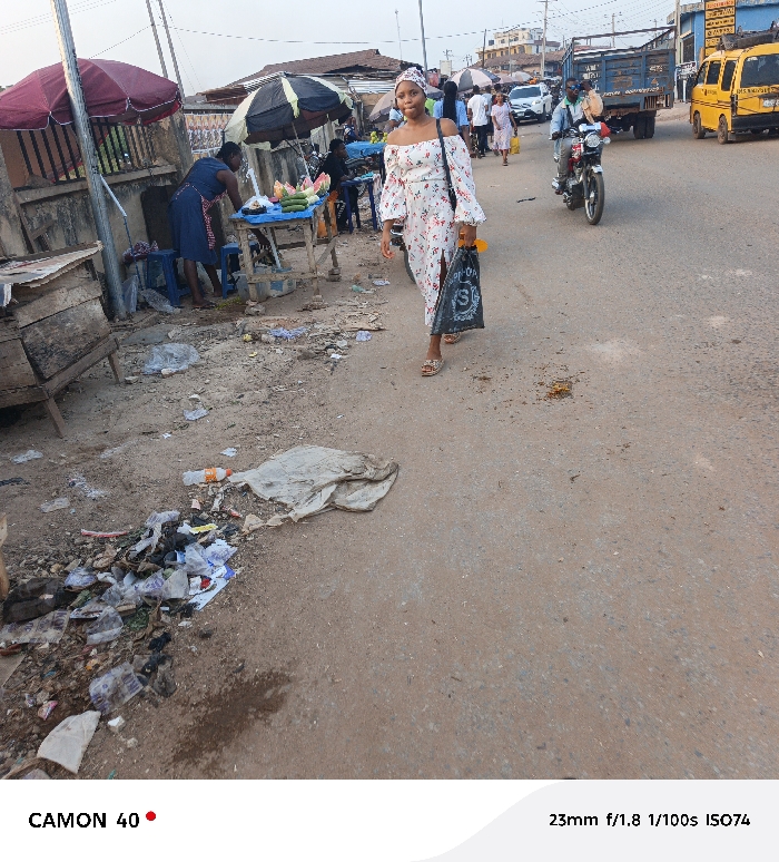 The image captures an outdoor street scene in Agbani, Nigeria, likely during late afternoon or early evening. In the foreground, a young woman walks along a dirt and paved road, slightly smiling at the camera. She wears a white, off-shoulder dress with a floral pattern and a matching headwrap, holding a black plastic bag with a prominent "S" logo. Her feet are visible in sandals.
To her left, a market stall is set up under a dark umbrella, with a woman in a blue dress attending to fruits and vegetables, including watermelons and cucumbers, displayed on a wooden table. Another red umbrella is visible further back. The ground near the market stalls and along the roadside is littered with debris, plastic waste, and dirt.
In the mid-ground and background, the street continues with ongoing activity. A man on a motorcycle drives past the main subject, and other pedestrians are visible further down the road. Several vehicles populate the street, including a yellow minibus (characteristic of public transport in Nigeria) and a blue truck. Buildings line the street, suggesting a commercial or residential area. The lighting is soft, indicating either early morning or late afternoon.
Visible text includes "CAMON 40" and "23mm f/1.8 1/100s ISO74" at the bottom of the image, detailing the camera model and settings. The black bag held by the main subject also shows an "S" logo.
