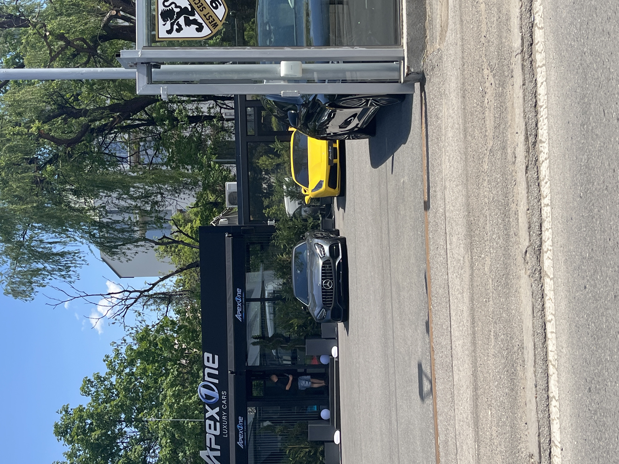 The image shows the exterior of "Apex One Luxury Cars" dealership in Sofia, Bulgaria. The building has a black facade with the company logo prominently displayed. Three luxury cars are parked outside: a gray Mercedes-AMG GT, a yellow Lamborghini, and a black car (likely another luxury model). A person is standing inside the dealership near the entrance. The street in front of the dealership is paved and includes a painted curb. Trees are visible in the background, partially obscuring the sky and surrounding buildings. A sign with the words "West Security" is also visible. The weather appears to be sunny.