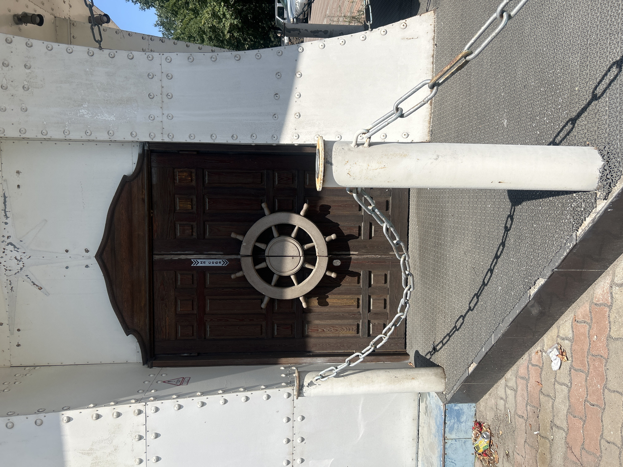 The image depicts a weathered wooden door with a ship's wheel decoration, set within a white, riveted structure. The door is flanked by two cylindrical white posts connected by a heavy metal chain. The ground in front of the door is covered with a grey textured mat, transitioning to a brick-like pathway to the left, where litter including a crushed can and some leaves are scattered. The structure appears to be an entrance to a building or an exhibit. The scene is illuminated by bright sunlight, casting distinct shadows. The architectural style suggests a maritime or themed establishment. This scene is located in Almaty, Kazakhstan.
