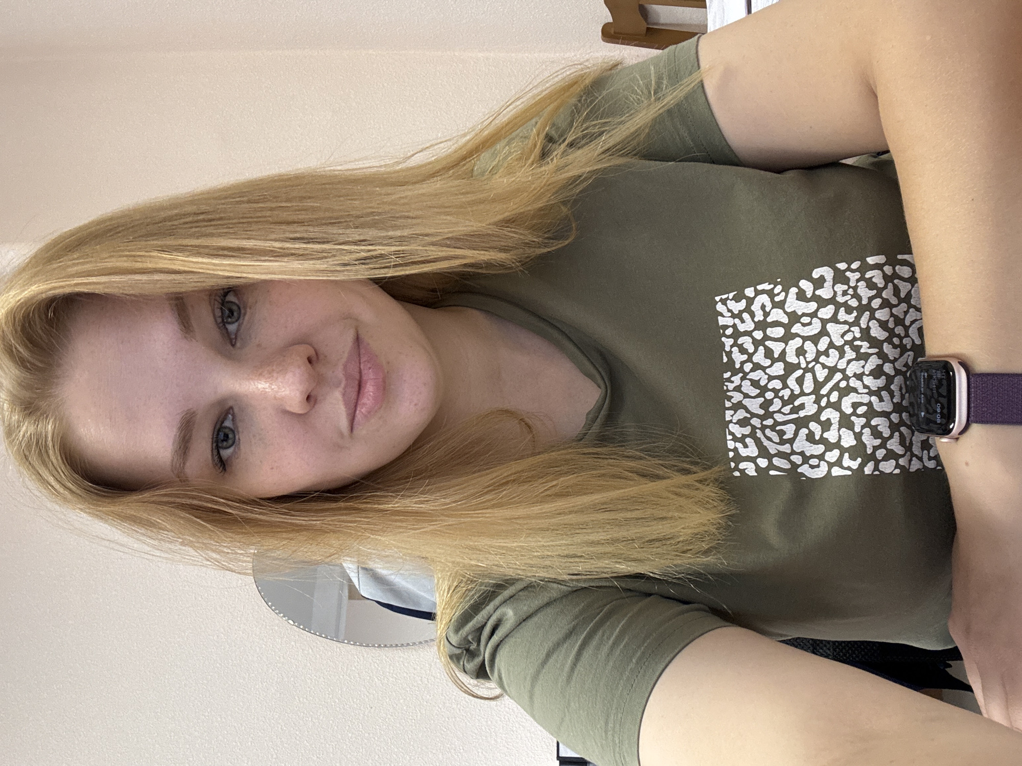 A young woman with shoulder-length blonde hair and fair skin is seated at a desk, looking directly at the camera. She is wearing a green t-shirt with a white, abstract pattern. Her left arm is crossed over her right arm, and she is wearing a purple watch on her left wrist. In the background, a wall with a textured finish is visible, along with a round mirror reflecting a portion of the room. To the right, a wooden chair is partially visible. The lighting suggests an indoor setting, possibly an office or a home study. The image does not provide any specific indicators of its location within the city of Заволжье, Russia, beyond the fact that it was taken there.