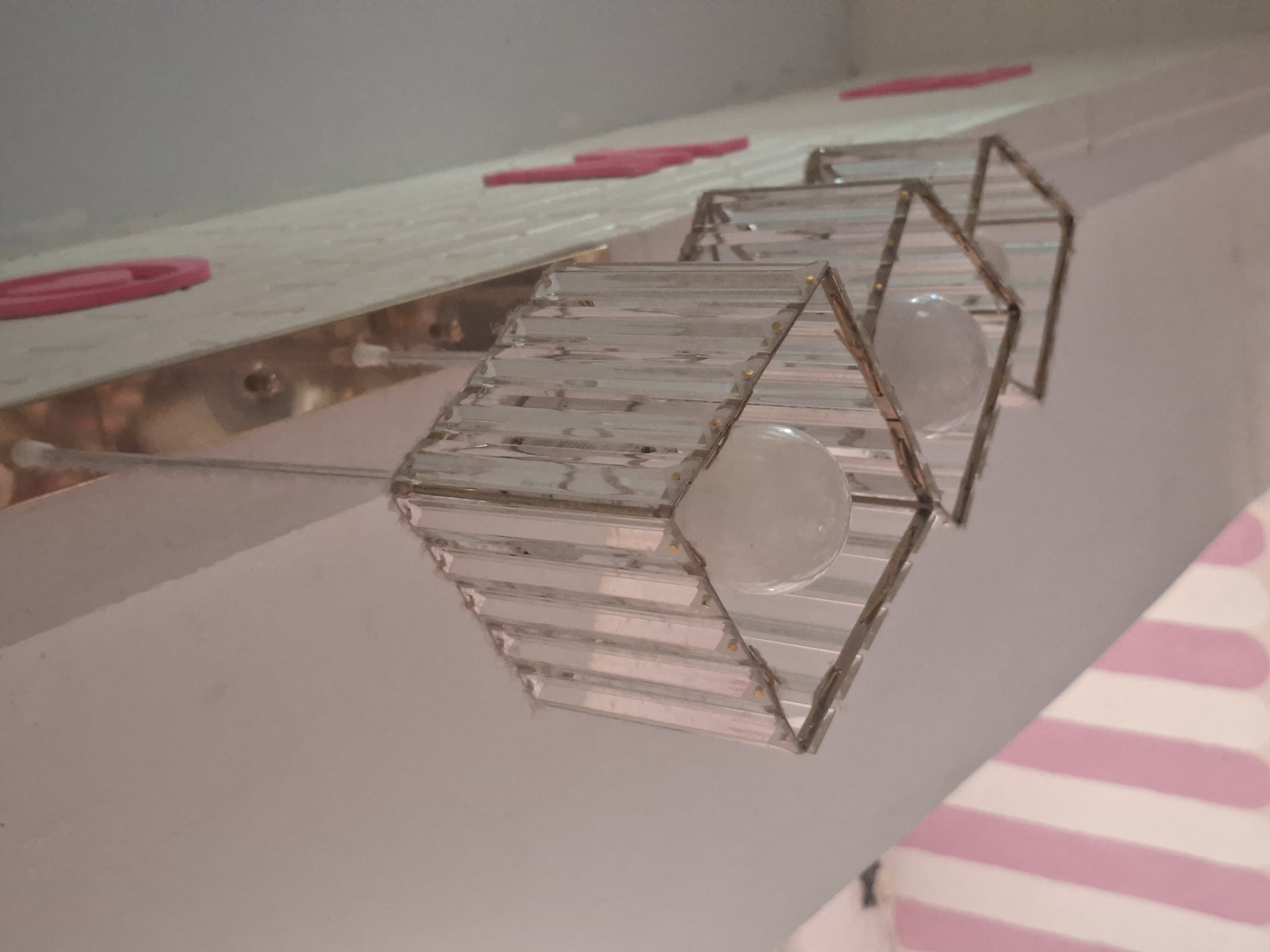 The image displays an upward perspective of three decorative light fixtures and their surrounding environment in Ciseeng, Indonesia. No people are visible.
The primary subjects are three hexagonal light fixtures, each constructed from clear, narrow, rectangular panels or strips, possibly glass or acrylic, held together by a metallic, likely brass-colored, frame. Each fixture houses an illuminated, spherical, frosted white light bulb. These fixtures appear to be suspended from a long, shiny golden or brass-colored strip that is affixed to a white or light-colored ceiling or upper wall.
Scattered on this golden strip and a surface just above it are several small, elongated, and slightly curved pink objects, possibly decorative elements or small plastic items. A distinct pink oval shape is visible to the left. The general setting suggests an indoor or covered outdoor space, possibly a commercial establishment like a cafe, restaurant, or event area, given the decorative nature of the lighting and pink accents.
In the bottom right corner of the image, a pattern of alternating pink and white or light cream stripes is partially visible, which could be part of an awning, tent canopy, or decorative textile. The illuminated bulbs suggest that the lights are on, implying either an indoor setting where artificial light is used or perhaps evening time. No specific activity is taking place, and no visible text is present in the image.