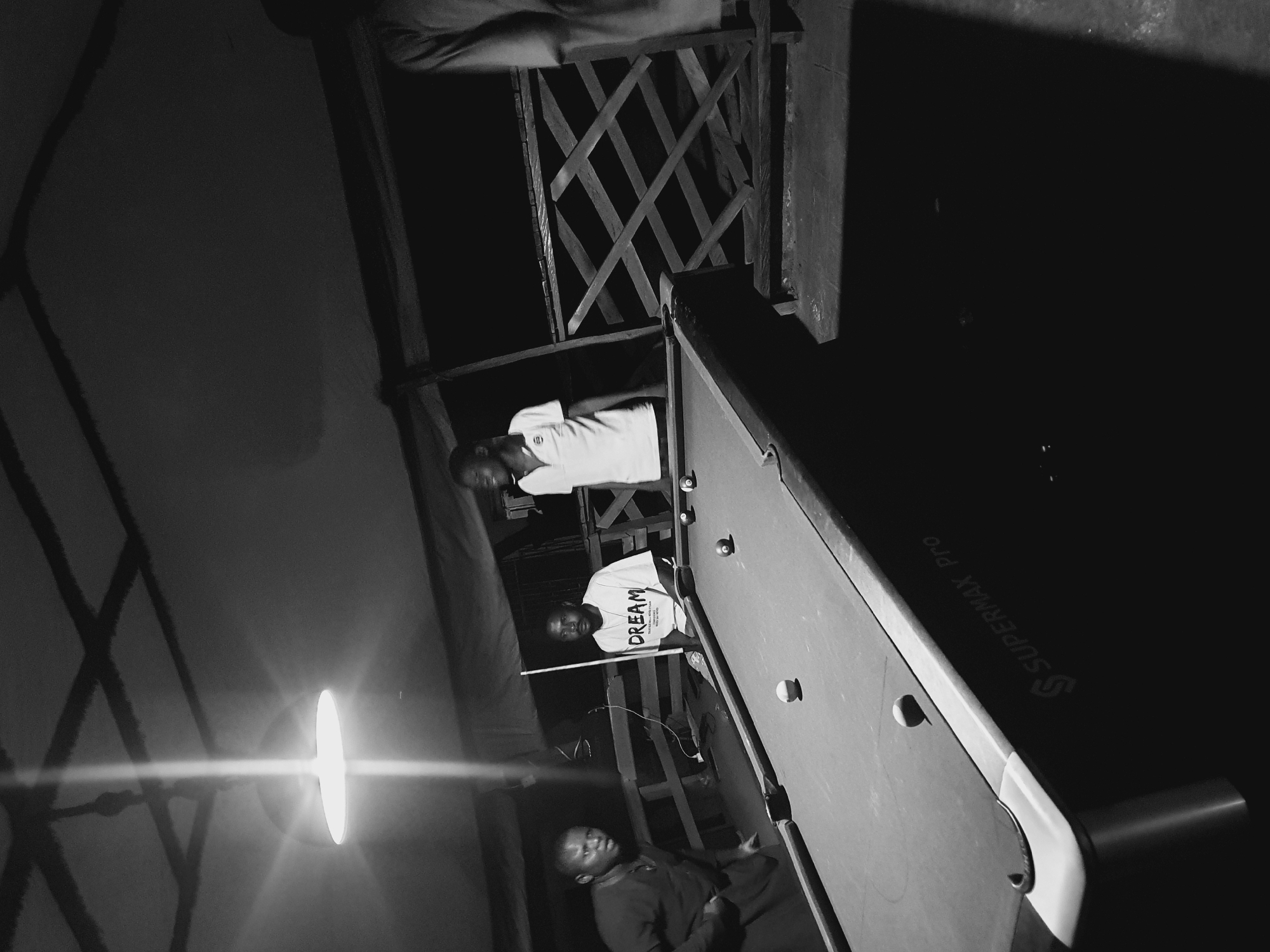 The image, taken in Lokoja, Nigeria, is a black and white photograph of a group of young men gathered around a pool table. The setting appears to be an outdoor or semi-outdoor area, possibly at night given the lighting and shadows. A bright, possibly artificial, light source is prominent on the left side of the frame, casting long shadows.
Three individuals are clearly visible. One person, wearing a white t-shirt with the word "DREAM" printed on it, is holding a pool cue and looking towards the camera. Another person, also in a white t-shirt, is standing slightly behind and to the left, looking away from the camera. A third person is seated or leaning near the pool table on the right, their face partially obscured and looking down.
The pool table itself is a central object, with several balls visible on its surface. The table has text visible on its side: "SUPERMAX PRO". The overall mood seems relaxed, with the individuals engaged in or observing a game of pool. The environment suggests a casual social gathering.