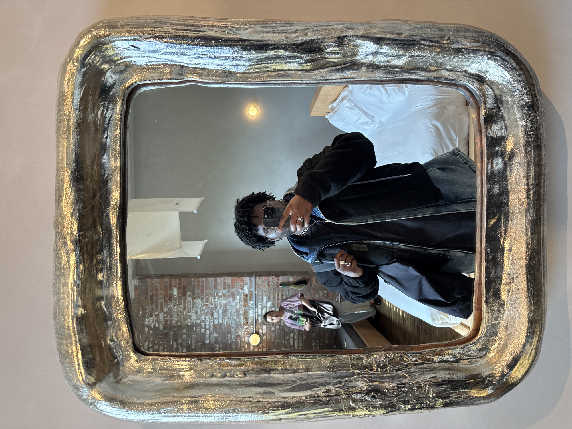 A person with dark skin and dreadlocks takes a mirror selfie in an indoor setting. They are wearing a dark jacket, a light-colored top, and possibly jeans, holding a smartphone in both hands to capture their reflection. Several rings are visible on their fingers.
The selfie is taken in a large, rectangular mirror featuring a thick, highly textured frame with a reflective silver finish, which appears metallic and has darker undertones. The reflections in the mirror reveal the surrounding room. To the right of the person taking the selfie, a neatly made bed with white bedding is visible. To the left, an exposed brick wall provides a rustic backdrop. Another person with short dark hair, wearing a lighter-colored top, is partially seen lounging or sitting against this brick wall. A warm-toned, circular light fixture is reflected on the ceiling within the mirror's view.
The scene suggests a casual moment captured in a well-lit interior space, possibly a bedroom or living area. The combination of the modern phone, casual attire, and the exposed brick wall hints at a contemporary or industrial-chic aesthetic, likely within a residence or hotel in the City of Hudson, United States. No discernible text is visible in the image.