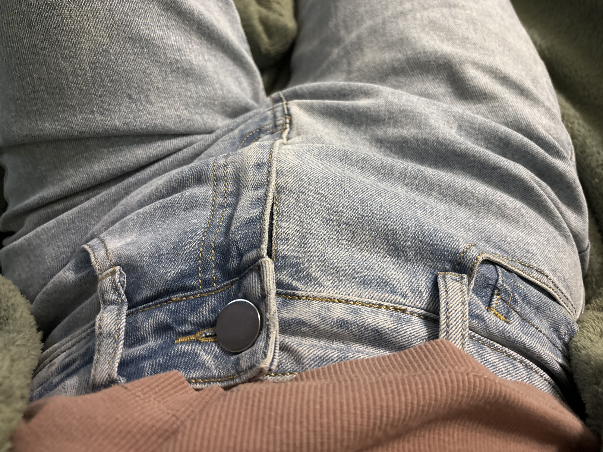 The image is a close-up, overhead shot of a person's lower torso, specifically their lap area, wearing light blue denim jeans. The jeans have visible stitching and a silver button at the waistband. A portion of a pink garment is visible at the bottom of the frame, peeking out from beneath the jeans. A green, textured fabric is visible on the left and right sides of the frame, suggesting the person is lying down or sitting on a soft surface. The scene is indoors, with neutral lighting. There is no indication of the location being in the city of Unknown, Russia.