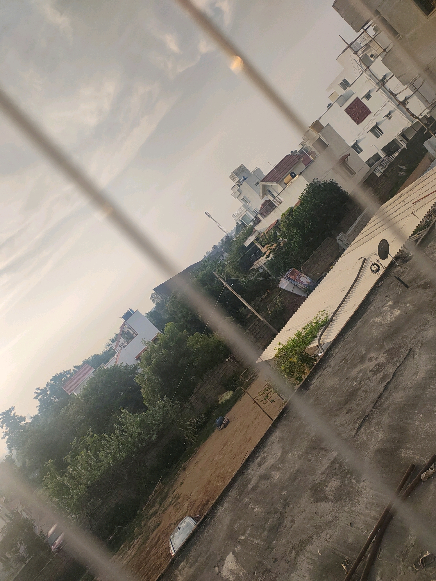 The image shows a view of the city of Kommasandra, India, through a wire fence. The perspective is from an elevated position, looking down and to the side. The sky is overcast with a pale, hazy light.
In the foreground and midground, there are several buildings, mostly white with some red-tiled roofs. These appear to be multi-story residential structures, some with balconies and windows. There are also trees and vegetation, with dense green foliage visible. The terrain seems uneven, with some dirt patches and paved areas.
On a rooftop in the midground, there is a satellite dish and some coiled cable. Another rooftop is covered with white corrugated material. In the lower section of the image, there is a dirt area with what appears to be a damaged or partially visible car. Further in the distance, more buildings and trees are scattered across the landscape. The overall impression is of a residential area within a city, with evidence of construction and everyday life.