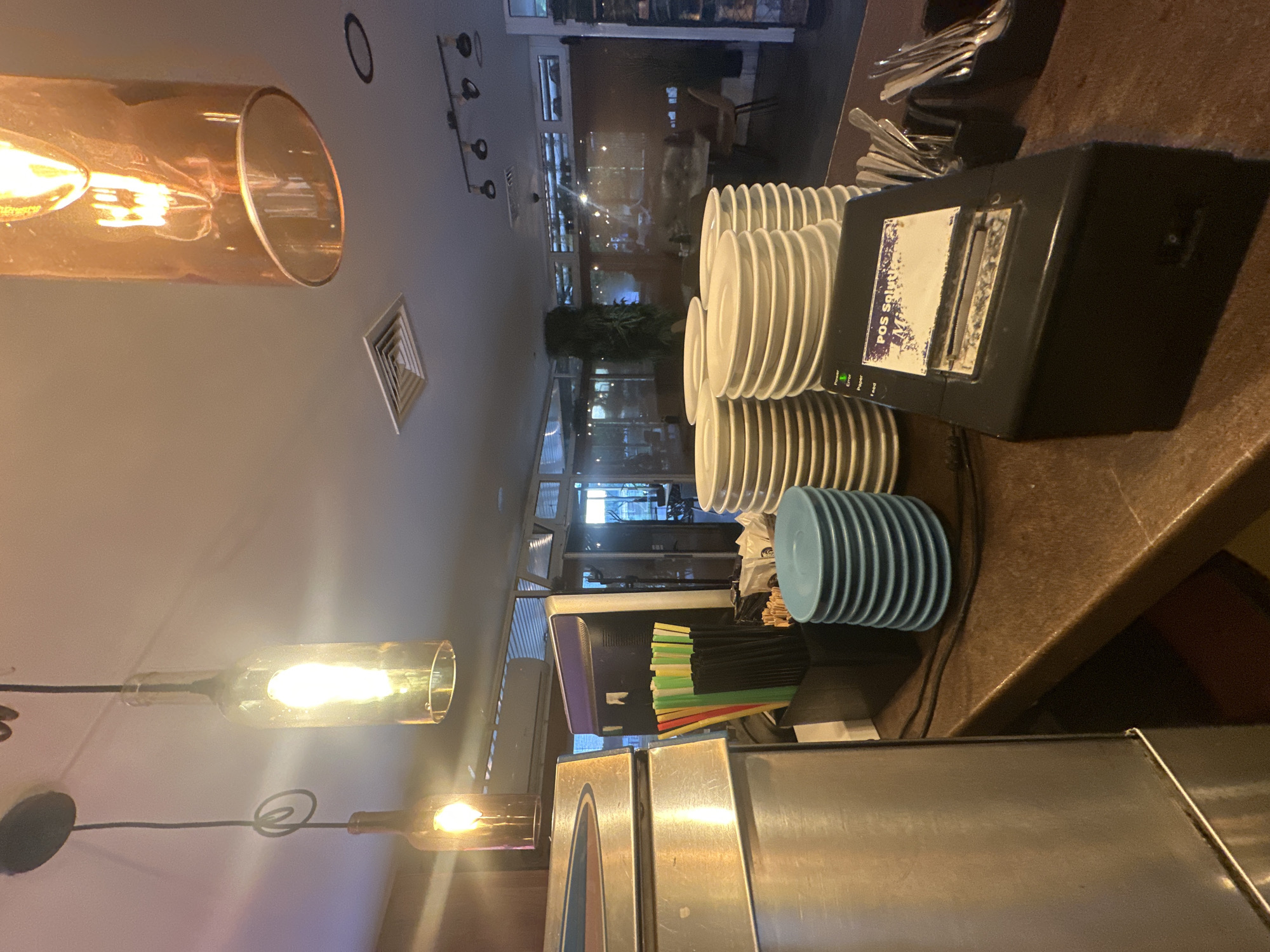 This image captures the interior of a commercial establishment, likely a restaurant or cafe, focusing on a service counter. The scene appears to be set indoors, with artificial lighting indicating it could be evening or night.
On the right side of the frame, a dark, speckled countertop holds stacks of clean white plates of various sizes, with a smaller stack of light blue plates towards the front. A black Point of Sale (POS) printer sits on the counter; a label on it partially reads "POS Soluti," and a green "Power" light is visible, suggesting it's active. Behind the printer, a container holds numerous silver utensils, including forks and spoons. Further along the counter to the left, multiple containers are filled with colorful straws in shades of yellow, green, red, and black. In the lower foreground, a section of stainless steel equipment is present.
The background features large windows, through which indistinct reflections of the interior are visible. Beyond the counter, glimpses of indoor seating (tables and chairs) and some potted plants can be seen, suggesting a dining area. The ceiling is light-colored, equipped with several light fixtures, including recessed lights and a track light with multiple spotlights. Prominent in the foreground and midground are stylish pendant lights: one in a large amber-tinted glass tube and others crafted from clear glass bottles, each housing an exposed bulb, casting a warm glow. An air vent is also visible on the ceiling.
No people are present in the image, giving the impression of a functional space either before opening, after closing, or during a quiet period. The overall setting is consistent with a commercial dining establishment in a town or city, such as Lom, Bulgaria.
