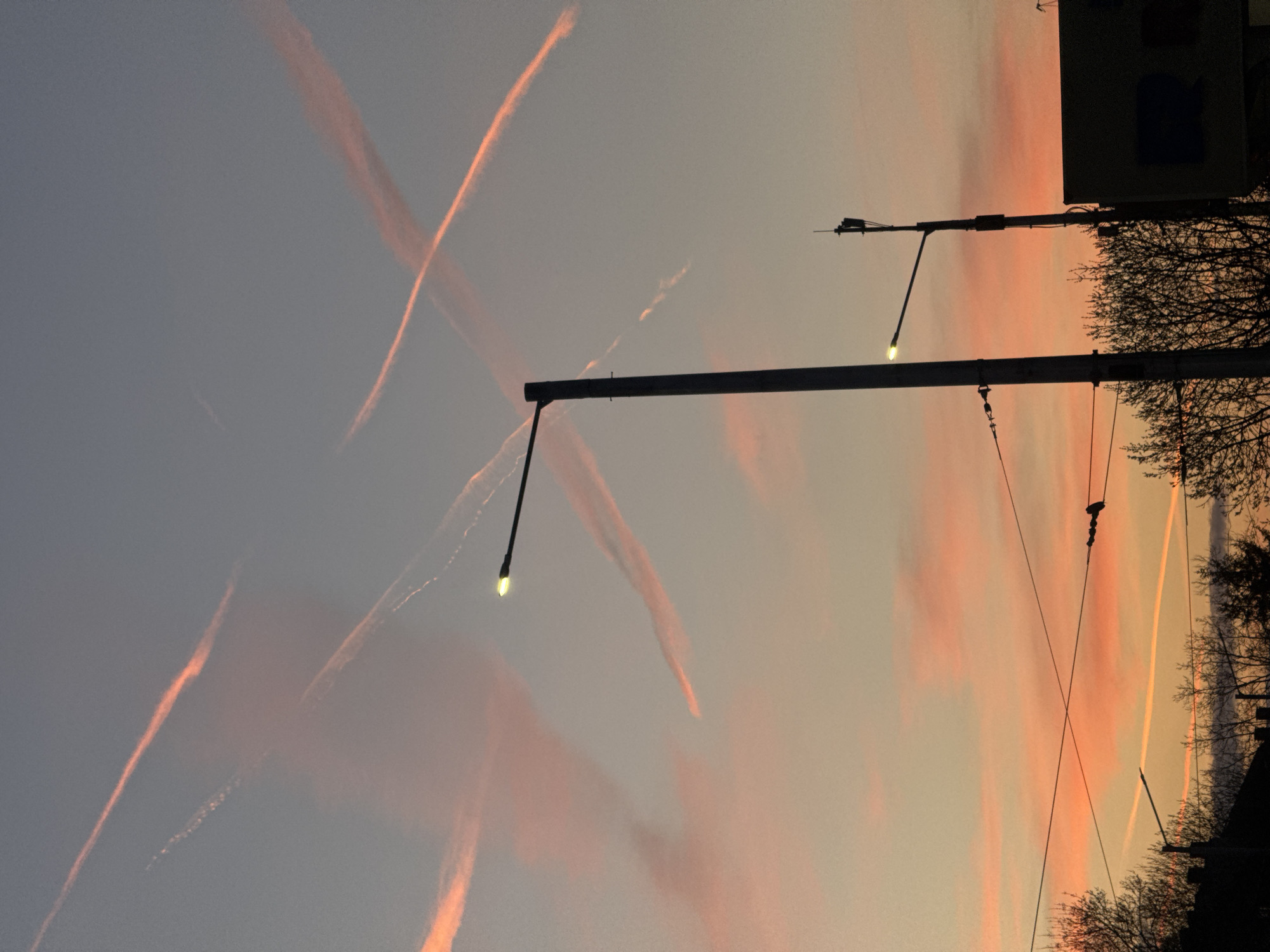 The image captures a dusk scene with intersecting contrails in the sky. A streetlight pole with two illuminated lights is centrally positioned. Electrical wires run diagonally across the frame, converging near the top. Silhouetted trees and a building edge are visible against the sunset sky on the right side. The location may be Sofia, Bulgaria, as inferred from the sky and streetlights. No people are visible.