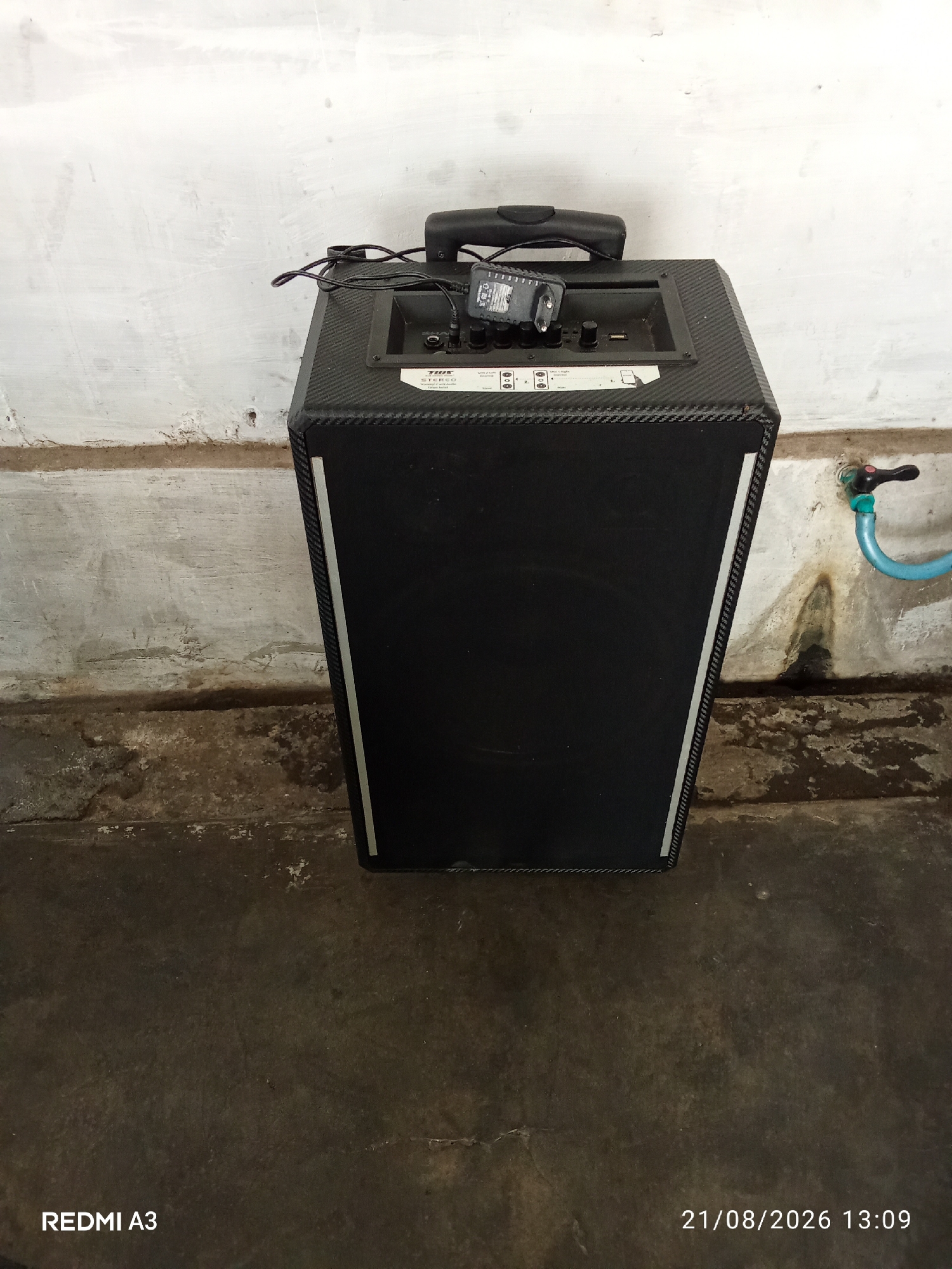 An outdoor, slightly low-angle full shot captures a black portable speaker with a carbon fiber-like texture, positioned in front of a weathered, off-white concrete wall. The speaker features a black retractable handle on top and a control panel with various buttons and ports. A black power adapter is plugged into one of the ports on the control panel. To the right of the speaker, a blue hose is connected to a faucet with a black and red handle, mounted on the wall. The ground in front of the speaker and wall is a dark, rough concrete surface. The lower left corner of the image displays "REDMI A3", and the lower right corner shows the date "21/08/2026" and time "13:09". The scene appears to be located in a utilitarian outdoor area, potentially a backyard or service area.