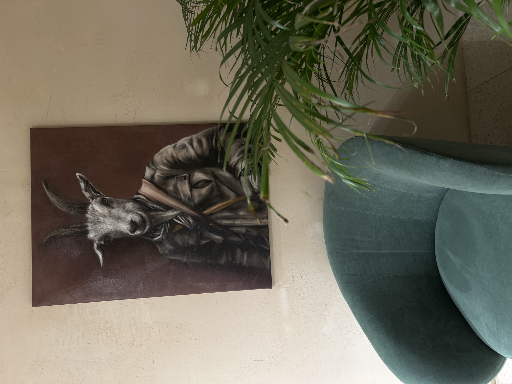 The image depicts a section of an interior wall, featuring a painting of a goat, a portion of a green chair, and foliage from a plant. The wall is a light beige or cream color with a textured surface.
The painting is rectangular and oriented horizontally. It is a depiction of a goat's head, rendered in shades of gray and brown. The background of the painting is a dark brown color. The goat has horns and seems to be holding something in its mouth.
To the right of the painting is part of a green chair. The chair appears to be made of a soft material, likely velvet or a similar fabric.
In the upper-right corner of the image is foliage from a plant. The leaves are long, thin, and green, suggesting a palm-like species.
Without additional information or context, it is impossible to determine if this scene is located in Saint Petersburg, Russia.