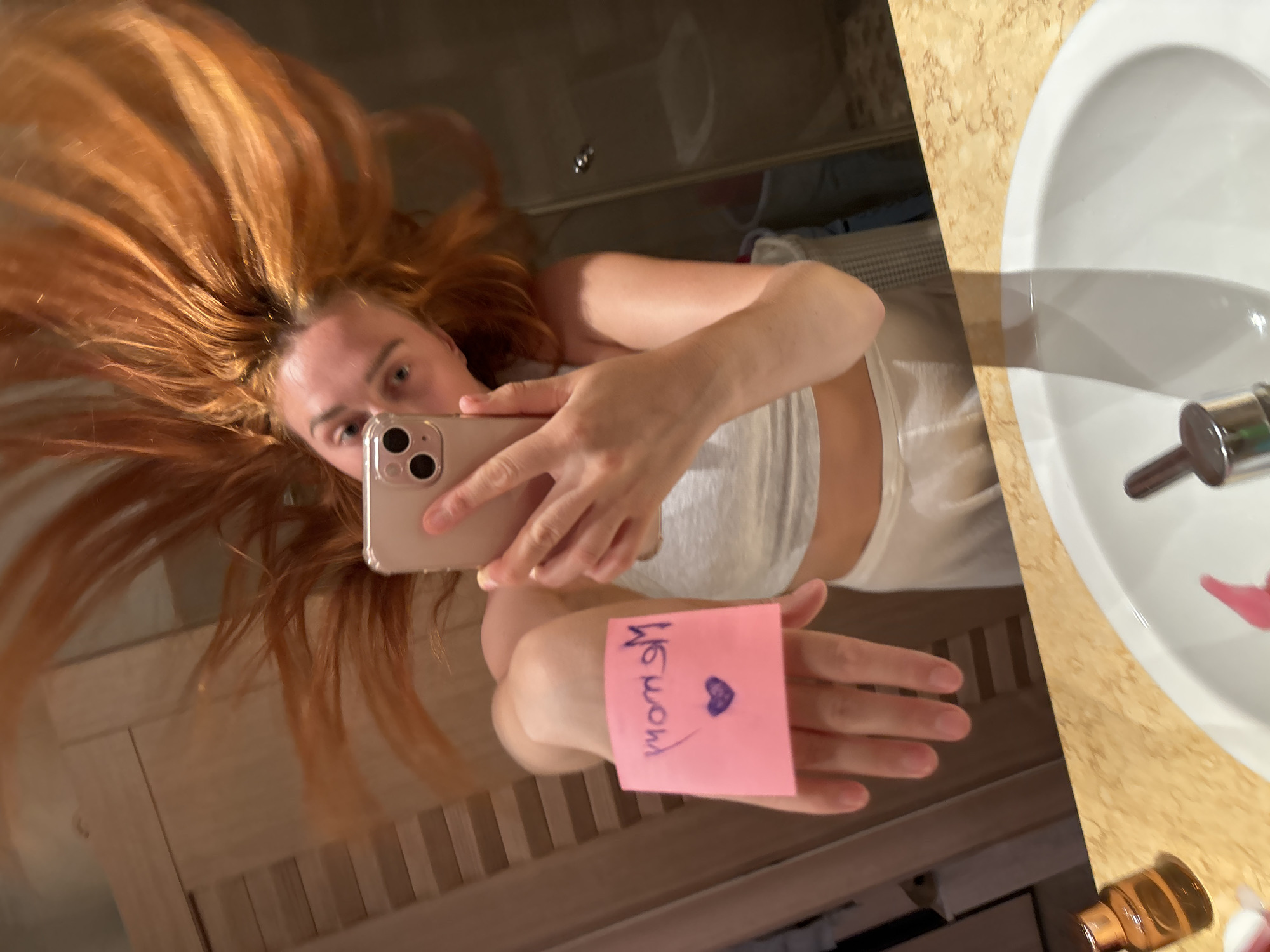 The image depicts a mirror selfie taken by a female individual in a bathroom in Budva, Montenegro. The subject has long, voluminous reddish-orange hair that appears styled upwards or in motion. She is wearing a light-colored crop top and bottoms. She holds a beige-colored smartphone in her right hand (viewer's left) to capture her reflection. Her left hand (viewer's right) is extended forward, displaying a pink square sticky note attached to her palm. The note bears mirrored text written in purple ink, reading "Mmom" (intended as "Mom" or "Mommy"), with a purple heart symbol beneath it. The foreground shows a white oval ceramic sink with a chrome faucet, integrated into a beige marble or stone countertop. An amber glass bottle and a partially visible pink dispenser are on the countertop. The background reflected in the mirror includes a light-colored wooden slatted structure to the left and a darker, indistinct area to the right.