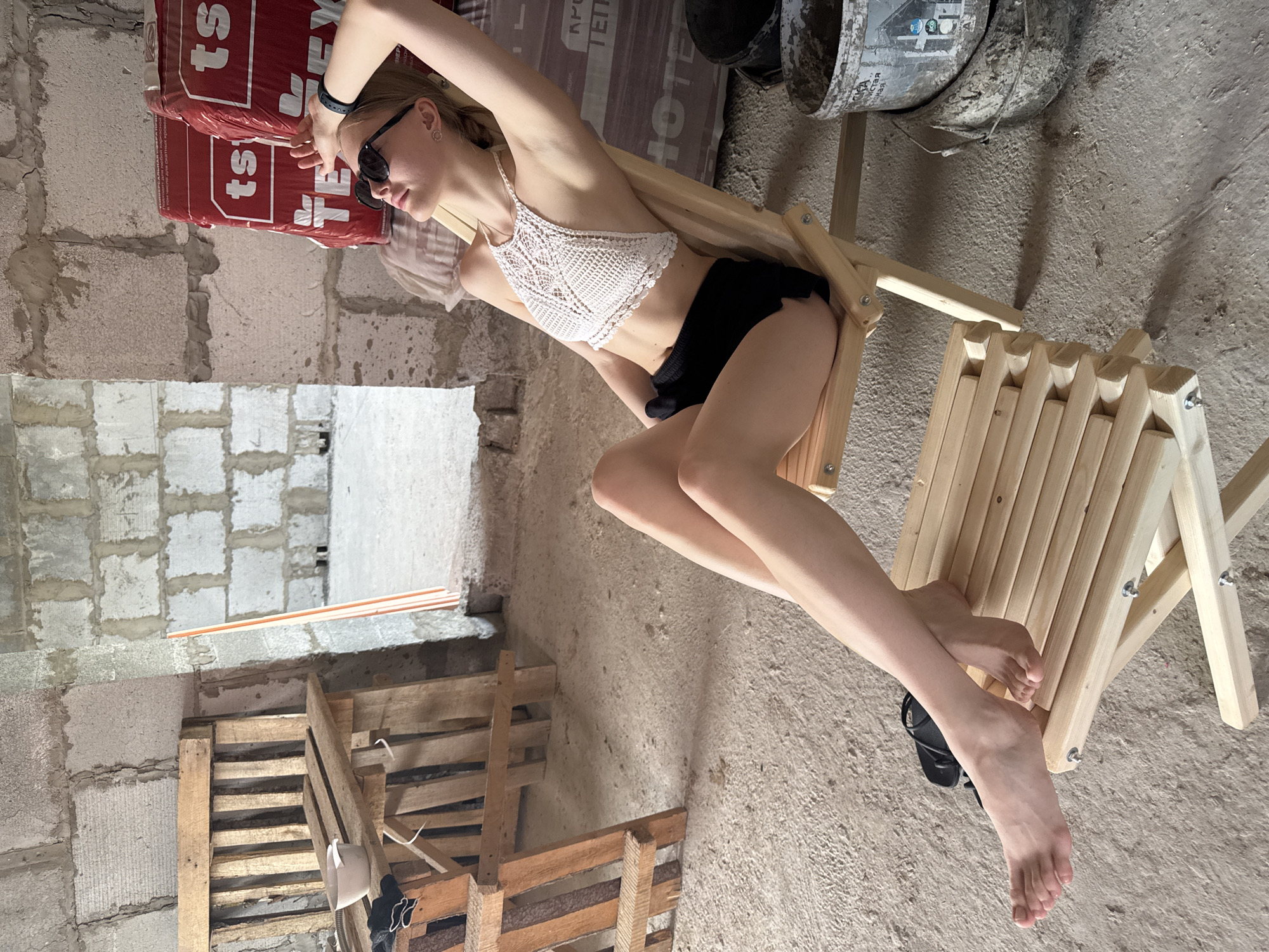 A young, light-skinned woman with blonde hair wearing sunglasses, a white crocheted top, and black shorts reclines on a light wood folding chair. She is indoors, amidst the rough, unfinished construction of a building.
Behind her is a stack of red-and-white construction materials and a doorway revealing an unfinished block wall. To her right is a dirty bucket on a wooden platform. Below her is a pile of wood. The flooring is unfinished concrete.
The location is in Perm, Russia.