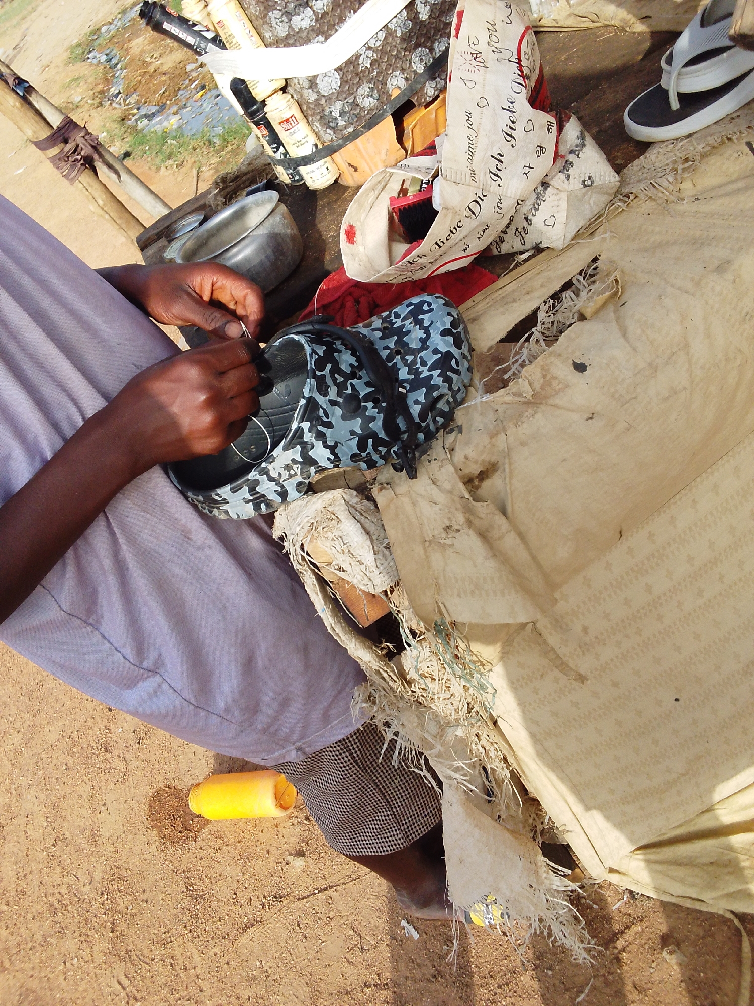 The photograph depicts a person mending a blue camouflage pattern Croc shoe with a needle and thread. The person is seated, wearing a light purple shirt and checkered brown and black pants. The mending is taking place on a makeshift surface consisting of a tattered beige cloth. Around the person and the shoe are various items including a metal bowl, some cans labeled "Shell", a bag with love-related German and Korean text printed on it, and a pair of white and black flip-flops. A yellow plastic container is on the ground near the person. The ground appears to be dry, sandy earth.
While not explicitly stated, the location is given as the city of Bauchi, Nigeria.