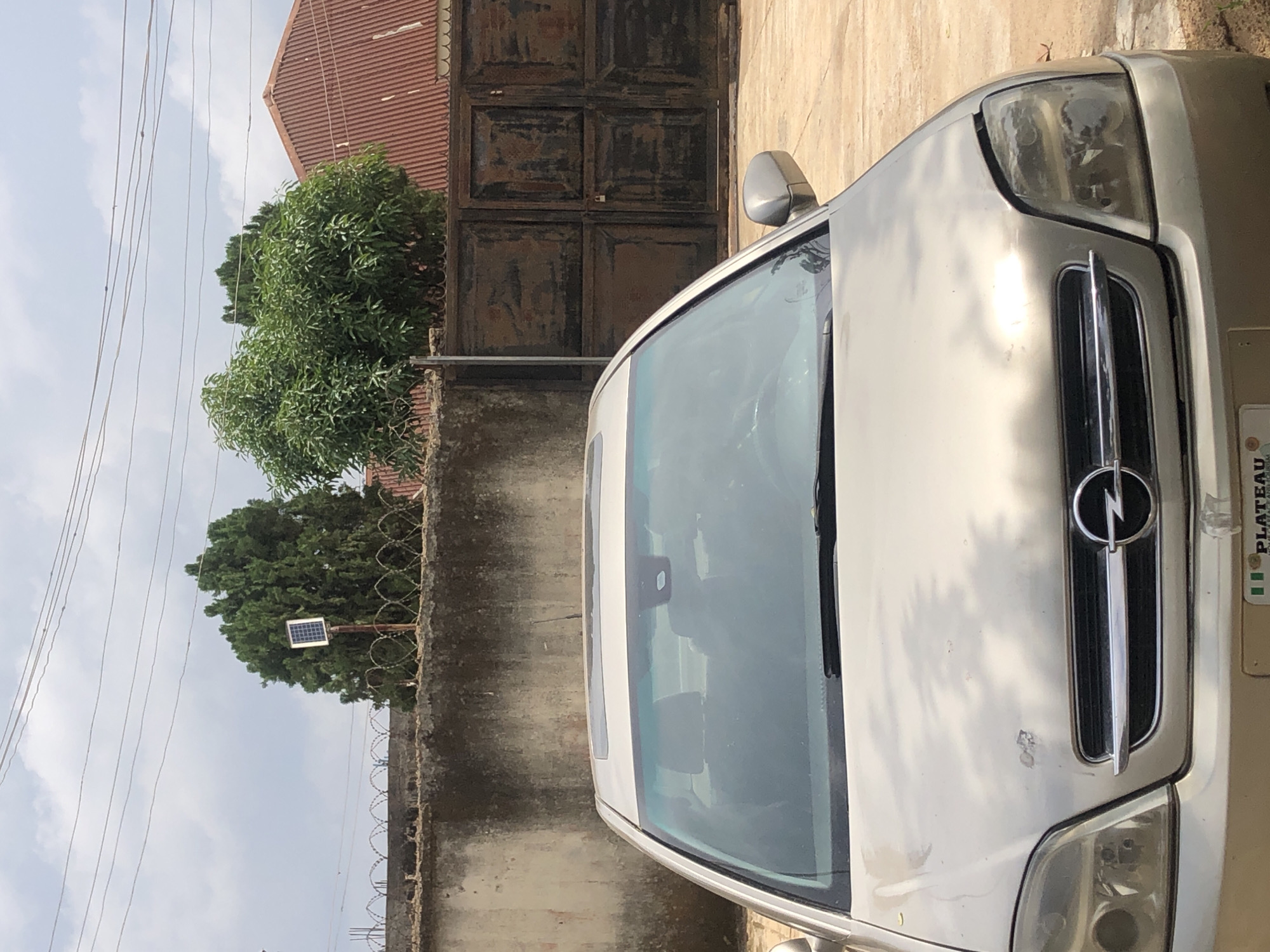 The image shows a silver Opel sedan parked in a yard. The car is facing the camera, and the hood and front bumper are visible. The car has a sunroof, and the windows are tinted. The car's headlights are slightly damaged. In the background, there is a concrete wall with barbed wire on top, and a large metal gate. Two trees are visible behind the wall, along with a building with a red corrugated iron roof. A solar panel is mounted on a pole behind the wall, near one of the trees. The sky is cloudy. The license plate of the car is partially visible and appears to have the text "PLATEAU" on it.