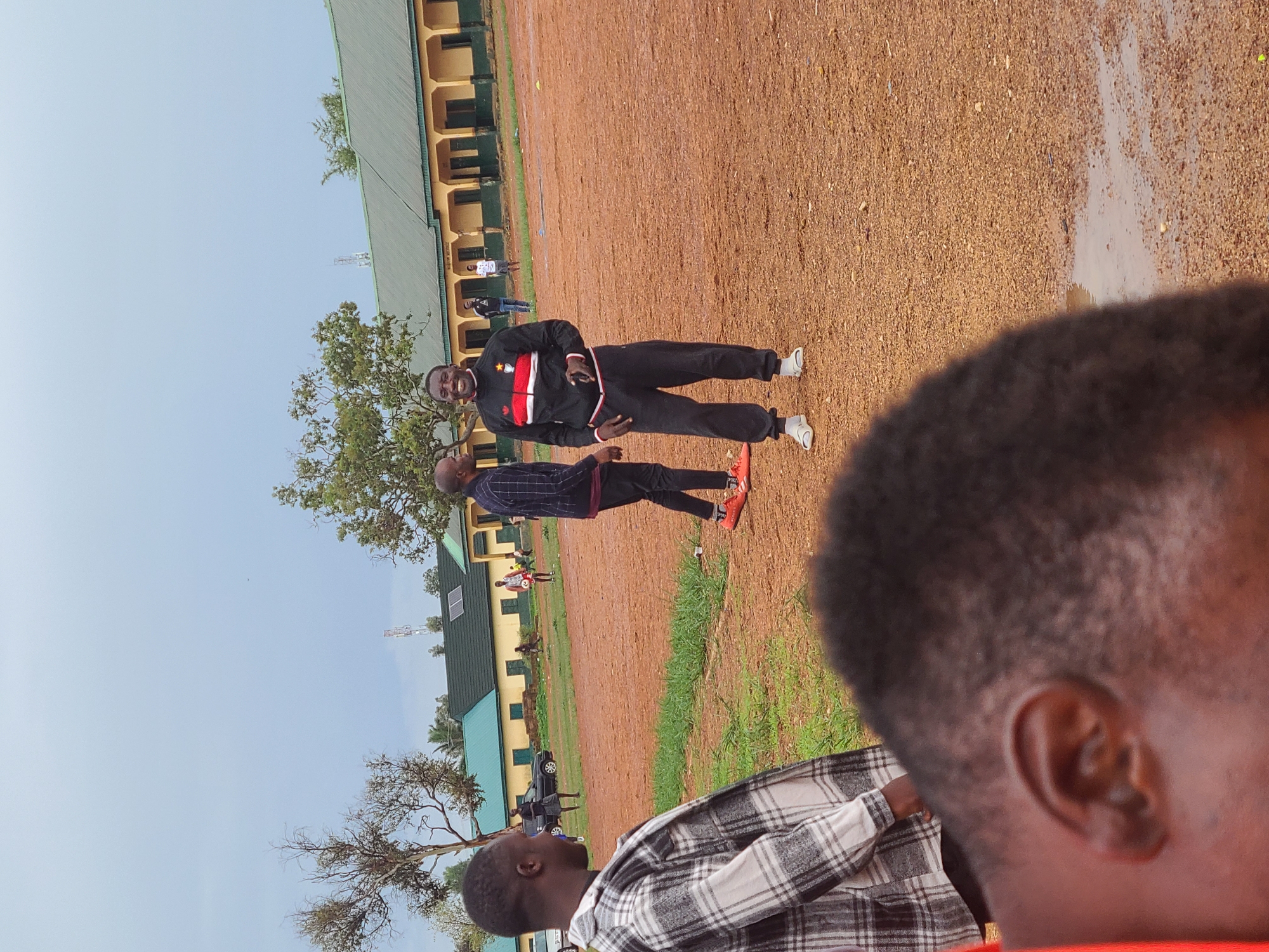 This daytime image, captured outdoors in Rafin Bauna, Nigeria, features a scene on a reddish-brown dirt and gravel field. In the immediate foreground, the right side of a person's head and ear is visible, with short dark hair, and they are wearing a black and white plaid shirt, seemingly observing the activity.
In the mid-ground, two men stand on the field. The man closer to the viewer is dressed in a dark, possibly plaid, long-sleeved shirt, dark pants, and red and black sneakers. He faces the central figure, a smiling man wearing a black tracksuit with prominent red and white stripes and a star-like emblem on the chest. This man is wearing white sneakers and appears to be looking towards the camera.
The background is dominated by a long, two-story building with a green corrugated metal roof and light-colored walls, featuring numerous windows or doorways. Several other individuals are scattered across the field and near the buildings, including one person in a white shirt and dark pants. A dark-colored car is partially visible near the structures. Trees with varying degrees of foliage are present across the landscape. A distinct wet patch or puddle is visible on the dirt field, indicating recent rain or standing water. A communication tower can be seen on the distant horizon against a clear, bright sky. The scene depicts a casual outdoor gathering or meeting. No text is visible in the image.