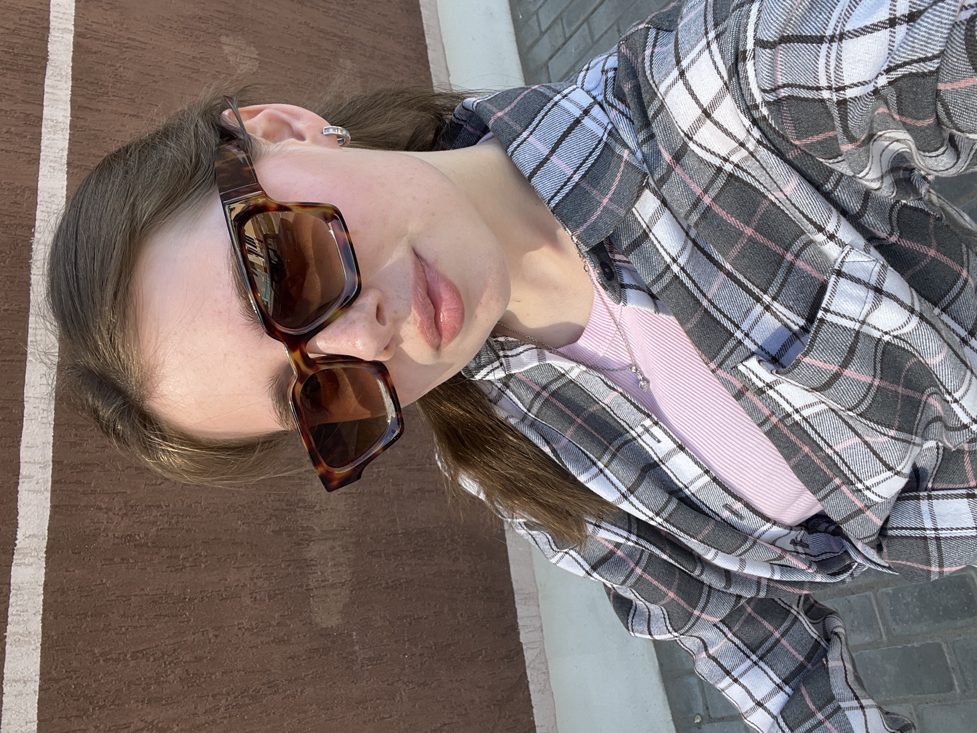 A young woman is posing for a selfie outdoors. She is wearing large tortoise-shell sunglasses, a gray and white plaid flannel shirt over a light pink top, and a silver hoop earring in her left ear. The woman is standing outside on what appears to be a brown brick sidewalk adjacent to a lighter brick or concrete surface. The photo angle is tilted, giving an overhead perspective. The location is near a pedestrian walkway in the city of Chaikovsky, Russia.