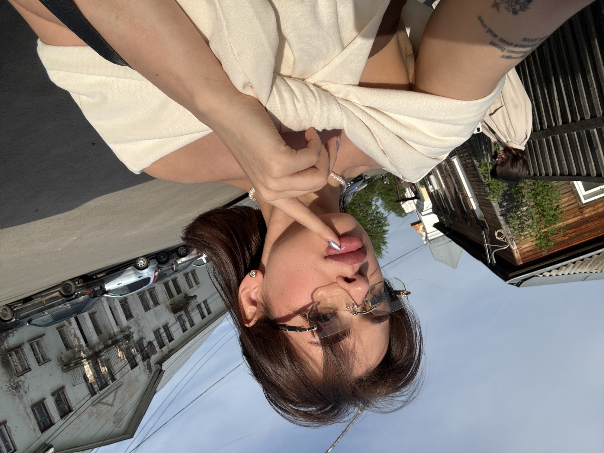 A woman poses outdoors in Sarapul, Russia. She wears glasses, a pearl necklace, and a light-colored top, placing her finger on her lip. The background includes buildings and cars lining the street, with a partly cloudy sky visible above. A fence and vegetation can also be seen on one side. The image is oriented upside down. The woman has a tattoo on her arm.