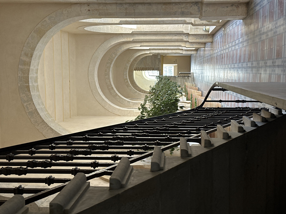 The image captures an arched corridor at the American Colony Hotel in Jaffa, Tel-Aviv, Israel. In the immediate foreground, a dark, ornate wrought-iron balustrade and the grey stone steps of a staircase ascend diagonally from the lower left, casting shadows. To the right of the staircase, a linear arrangement of potted, leafy green trees in terracotta planters extends along the left side of the long, receding corridor. The right side of the corridor is characterized by a series of prominent, light-colored stone arches supported by thick pillars, allowing natural light to illuminate the space. The floor features a consistent pattern of reddish-brown and lighter square tiles. The walls and ceilings are painted a light, uniform beige, with additional plastered archways visible deeper within the passage. In the background, the corridor ends with an arched window and a less distinct staircase. The scene is devoid of individuals, activities, or interactions.
