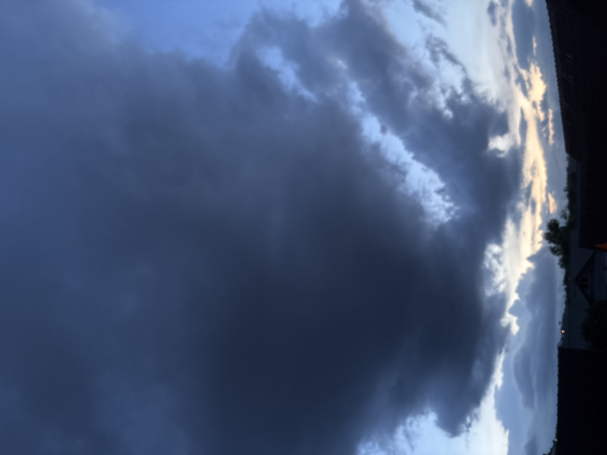The image depicts a sky with dark, heavy clouds. The majority of the frame is filled with a large, ominous cloud formation that appears to be a storm cloud. Patches of lighter blue sky can be seen peeking through the edges of the dark cloud cover. In the upper right corner, trees and a building are partially visible, indicating a residential area. The image is taken from a low angle, looking up at the sky. It is likely taken during the late afternoon or early evening. The scene is located in บ้านบางรักษ์, Thailand, based on the given location. The overall mood is one of impending rain or a storm.