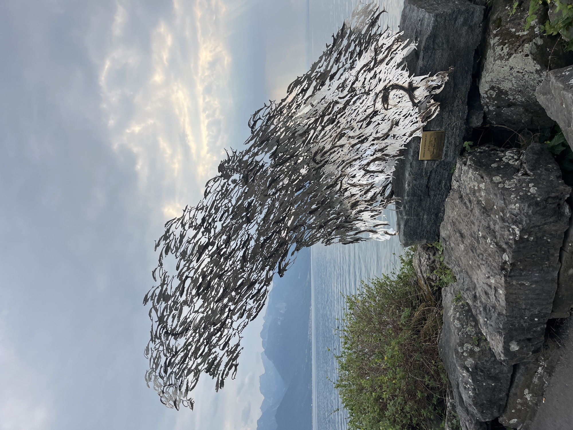 This image depicts an outdoor sculpture, identified by a plaque as "Above Sea Level" by Doug & Dave, which was part of the Biennale de Montreux 2017. The sculpture, made of numerous metallic fish-like forms, appears to be moving as a school or wave. It is positioned on a rocky embankment overlooking a large body of water, likely Lake Geneva, with mountains visible in the background. The sky is overcast with some rays of sunlight breaking through. In the foreground, there are large, grey stones and a green bush. The overall scene is serene and natural, with the sculpture serving as a striking focal point.