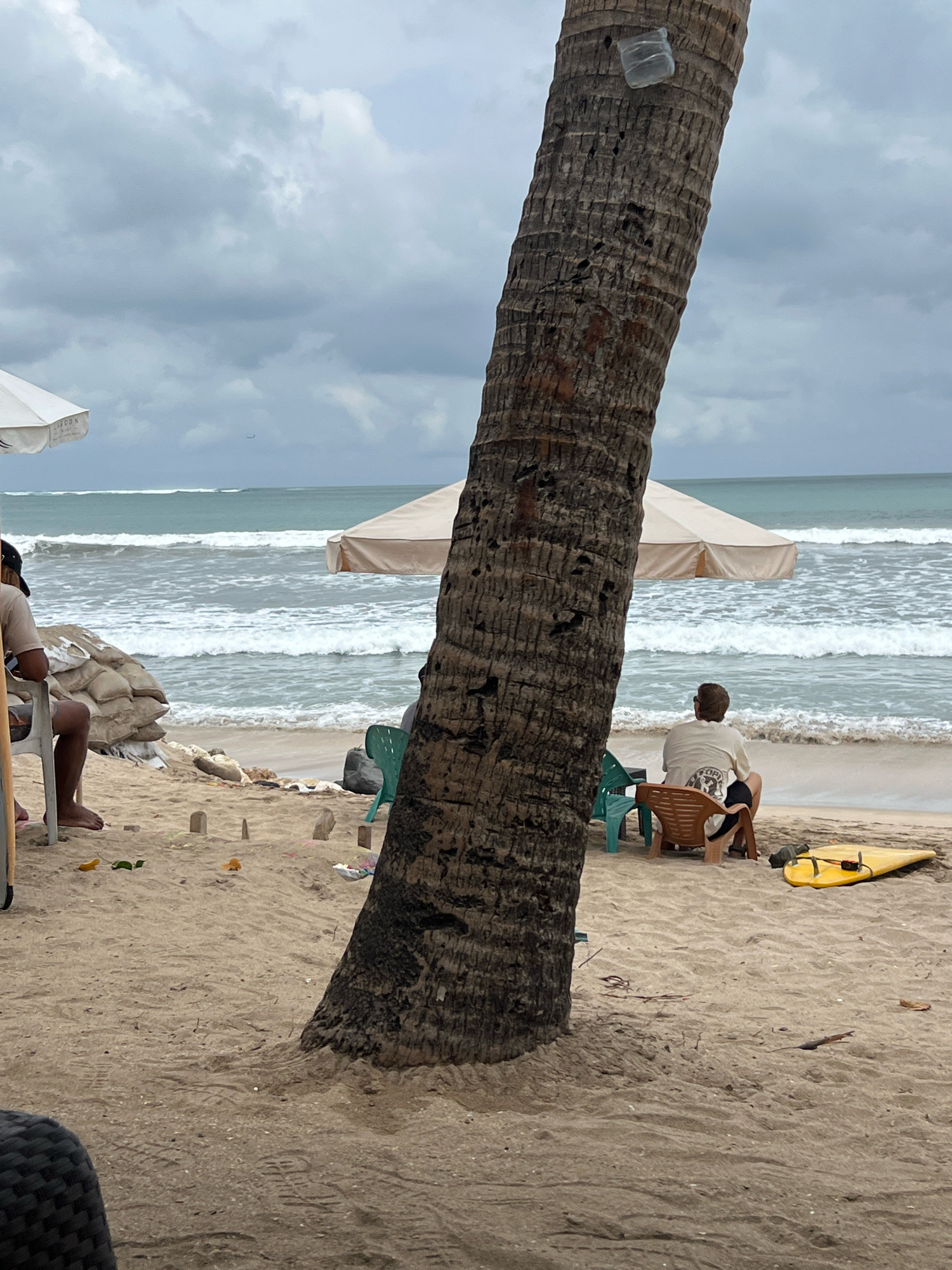 A medium shot captures a sandy beach in Kuta, Indonesia, under a cloudy sky. In the foreground, a large palm tree trunk dominates the left side of the frame. To the right of the palm tree, a person sits in a brown chair, facing away from the camera, looking out at the ocean. A yellow surfboard rests on the sand to their right. In the background, the ocean exhibits moderate waves, and two beach umbrellas provide shade on the sand. Sandbags are visible on the left side of the image. The overall scene depicts a relaxed beach setting with overcast weather.
