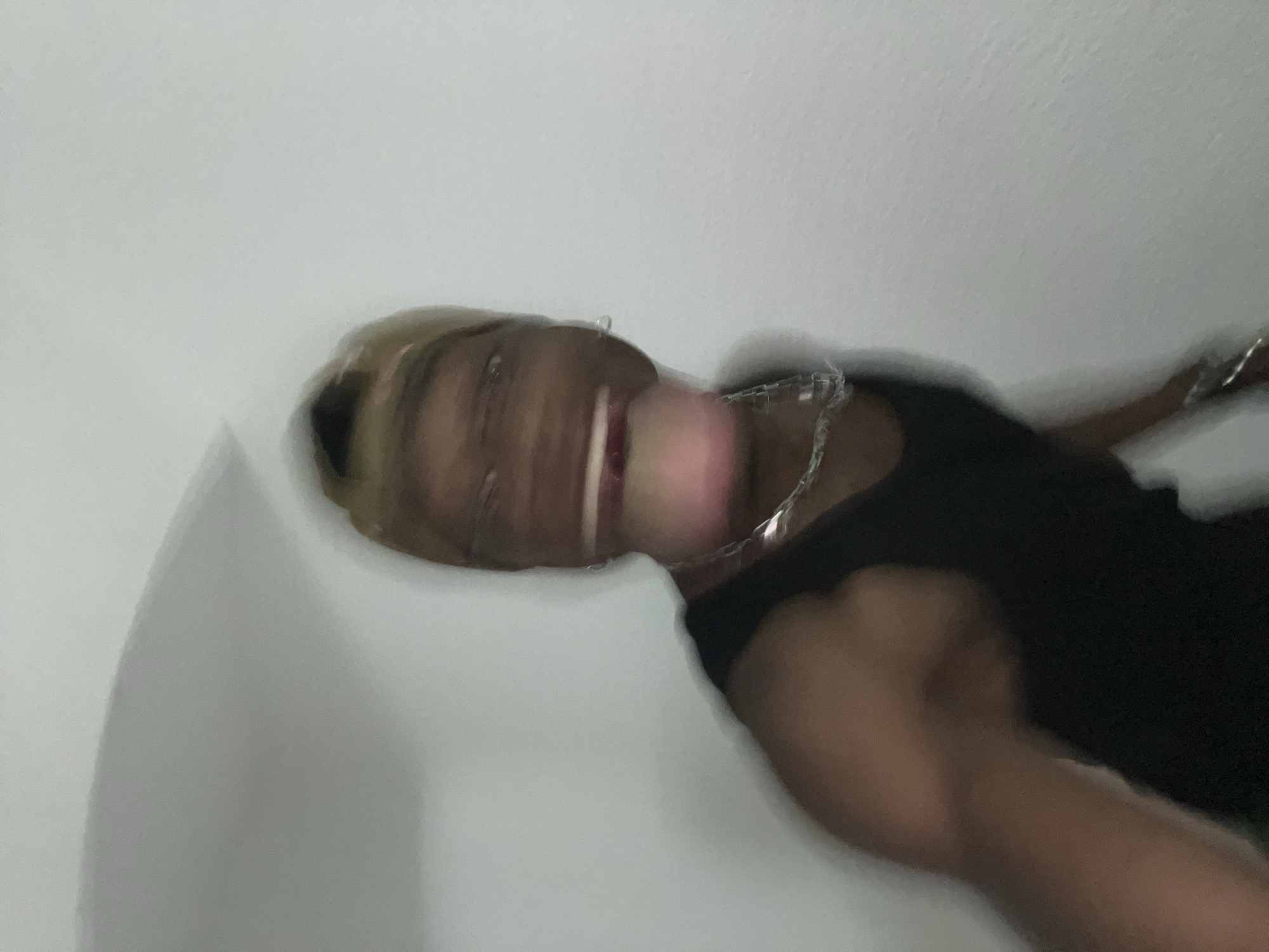 A highly blurry, low-angle photograph captures a person smiling broadly, seemingly looking down towards the camera. The subject has dark skin and what appears to be light-colored or blonde hair. They are wearing a dark, possibly black, tank top or sleeveless shirt, and a silver-colored chain or necklace is visible around their neck. The person's mouth is wide open in a smile, revealing teeth, and their face conveys an emotion of happiness or laughter. Due to the significant motion blur, facial features are indistinct, creating a stretched and smeared effect, especially around the mouth and eyes. The background is a plain, light-colored surface, likely a wall or ceiling, suggesting an indoor setting. No discernible text is visible in the image. The overall impression is a spontaneous, candid moment captured with movement.