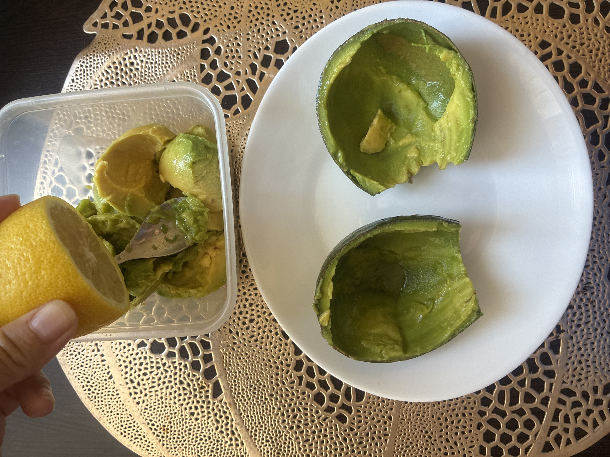 In a domestic setting, a person's hand is squeezing a halved lemon over a clear plastic container filled with scooped avocado flesh and a fork. The container rests on a gold-colored, textured placemat. In the foreground, a white plate holds two halves of an empty avocado skin, also appearing to have had some flesh scooped out. The environment is indoors, with a dark background, possibly a table. There is no discernible indication of the city of Sochi, Russia.