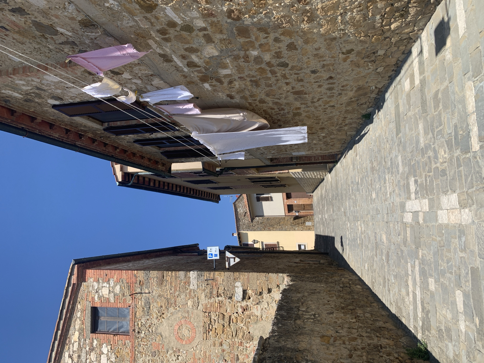 The image depicts a narrow cobblestone street in San Quirico d'Orcia, Italy. The street is lined with old stone buildings, some with exposed brickwork. Laundry is hanging from a clothesline strung across the street from one of the buildings. The sky is clear and blue, indicating a sunny day. The architecture suggests a historic Tuscan town. The scene is tranquil and shows signs of everyday life.