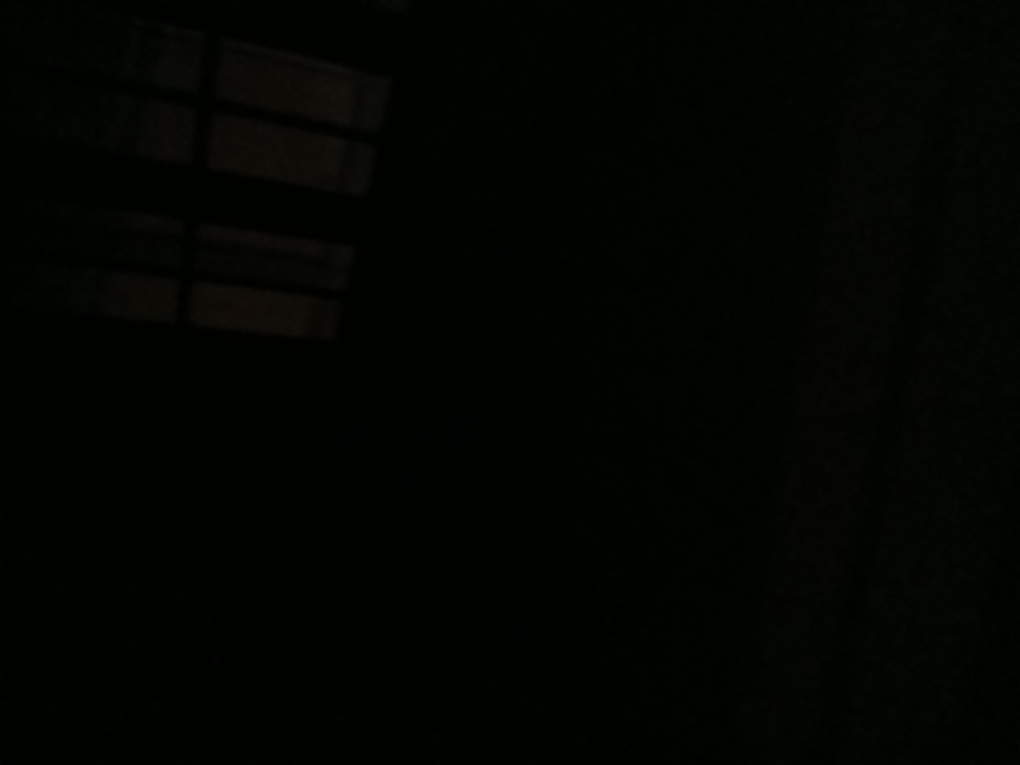 The image shows a dark, abstract scene. It appears to be a reflection on a dark surface, possibly a window at night. The reflection shows the faint outline of what looks like window panes with mullions, suggesting an interior space with lighting. The lower portion of the image is completely black, offering no discernible details. There are no people or identifiable objects present. The location in Tenggarang, Indonesia, is not evident from the visual information provided.