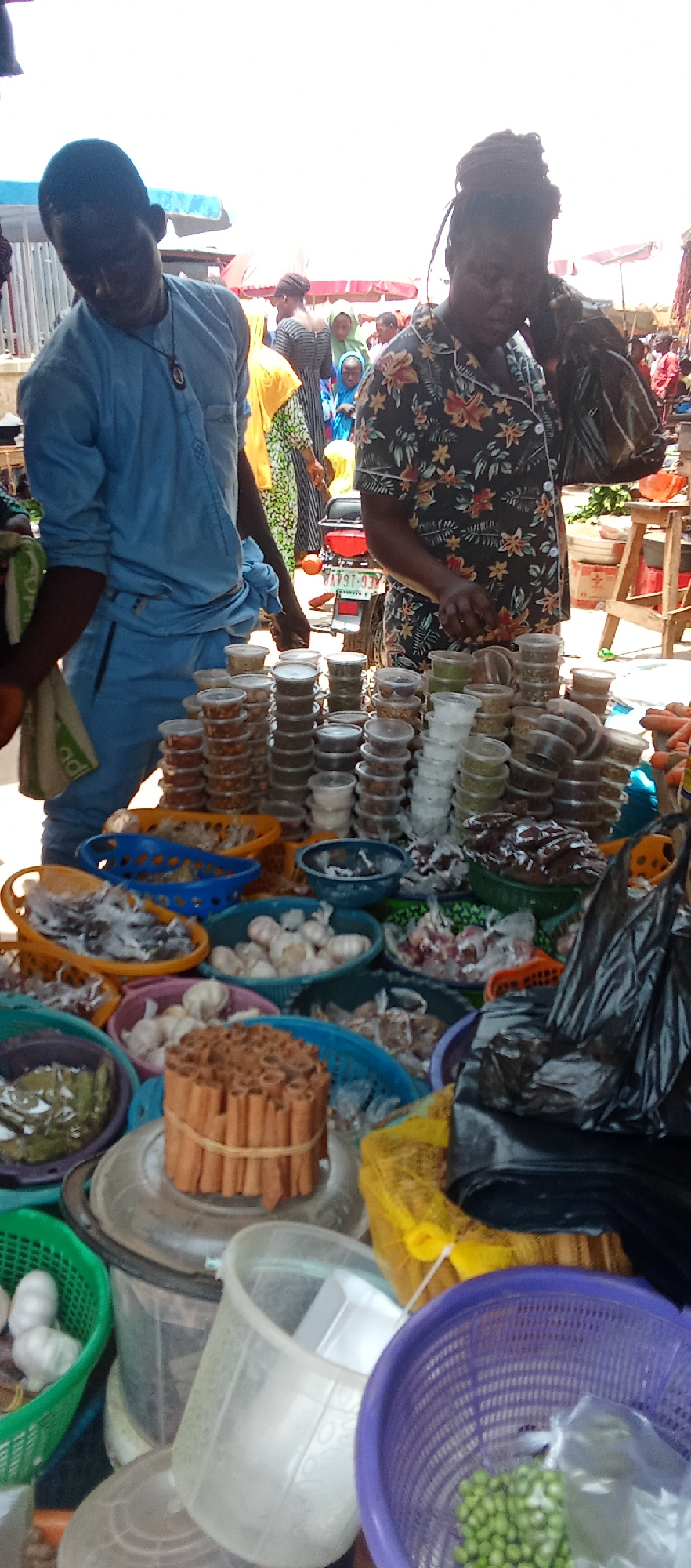 The image captures a vibrant market scene in Akwanga, Nigeria. Two individuals are prominent: a man in a light blue shirt and a woman in a dark floral-patterned shirt. They are standing near a display of goods, including stacks of small, clear containers filled with spices or ingredients. In front of them are various baskets filled with items such as garlic, cinnamon sticks, and other unidentified food products. In the background, there are other people, some covered up, going about their business in the busy market. A motorcycle is parked nearby. The market appears to be outdoors, and sunlight suggests it is daytime.