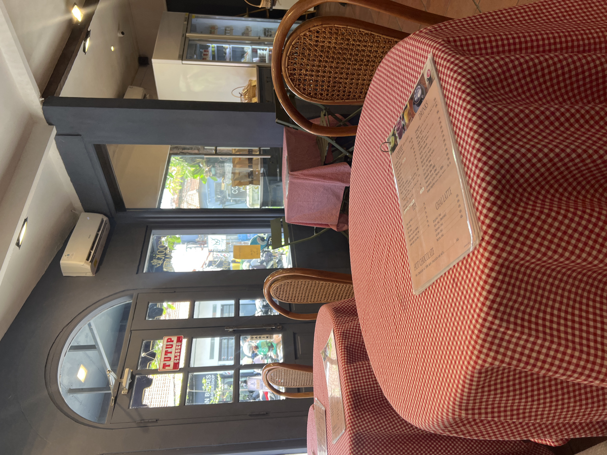 The image captures the interior of a café, seemingly in North Kuta, Indonesia. The focus is on tables covered with red and white checkered tablecloths. Brown rattan chairs are placed around the tables. A menu card stands on the table, listing various coffee and drink options. Outside the cafe is a partially visible storefront of "In Blades Barbershop". The door to the cafe has a "Tutup/Closed" sign on it. An air conditioning unit is mounted on the wall. The café appears to be located on a street with shops and some vehicular traffic. A refrigerated display cabinet is visible in the background.
