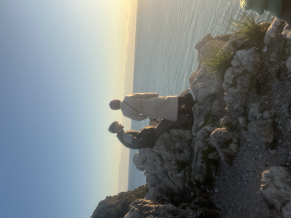 The image captures two individuals positioned on a rocky outcrop overlooking the sea in Alanya, Turkey. The scene is illuminated by the low-angle light of either sunrise or sunset, with the sky transitioning from clear blue to a warm golden hue at the horizon on the right.
In the midground, a male individual is seated on a large, grey rock. He wears a dark vest over a lighter long-sleeved garment and dark trousers, looking towards the right. To his right and slightly behind him, a female individual stands, primarily with her back to the viewer, facing the open sea. She is dressed in a light-colored, knee-length coat, dark legwear, and white footwear, with her head covered by a reddish-brown headscarf.
The immediate environment consists of rough, irregular grey rock formations and sparse clumps of green vegetation. A narrow dirt path is visible on the lower left. Beyond the individuals, the vast expanse of the Mediterranean Sea extends to the horizon, displaying a gently rippled surface. A steep, vegetated cliff face rises in the far left background.