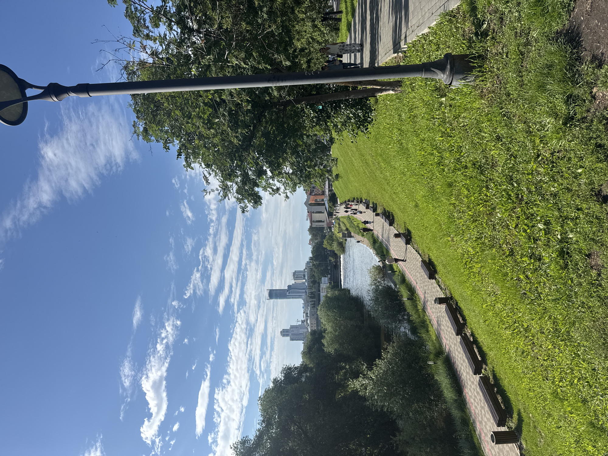 The image captures a scenic view of the Iset River embankment in Yekaterinburg, Russia. A paved walkway runs along the river, lined with benches and lampposts. Lush green grass covers the embankment, sloping upwards towards a dense canopy of trees on the right side of the frame. In the distance, modern skyscrapers and buildings rise above the treeline, indicating the city's urban landscape. Several people are visible walking along the path, enjoying the sunny weather and clear blue sky dotted with white clouds. The river itself appears calm, reflecting the surrounding greenery and buildings. The overall scene depicts a vibrant public space in Yekaterinburg, blending natural beauty with urban development.