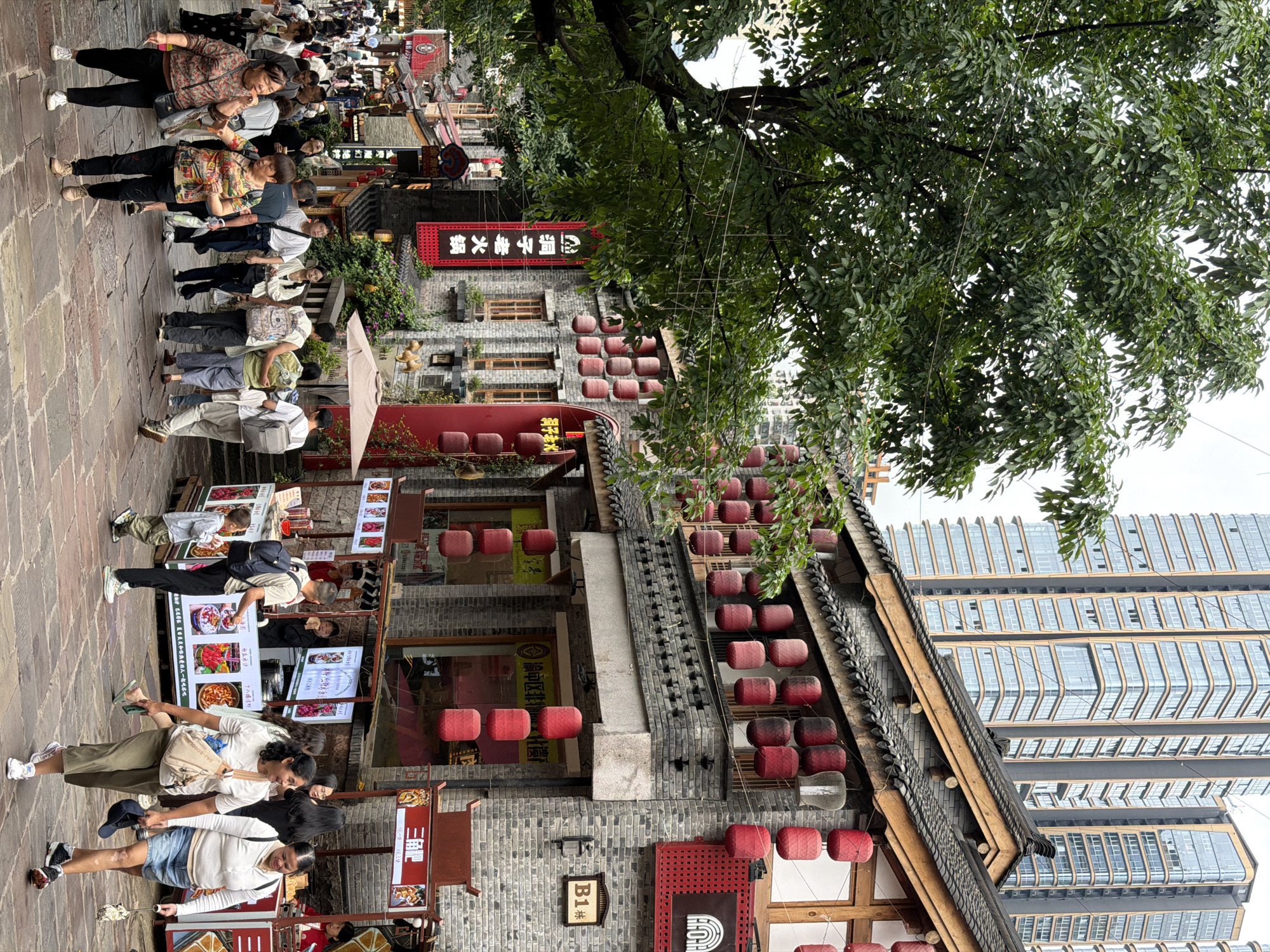 The image depicts a bustling street scene in Yuzhong District, China, characterized by traditional architecture adorned with red lanterns and contrasted by modern high-rise buildings in the background. A group of people are walking along a stone-paved street lined with shops and food stalls. The foreground shows several individuals, including two women in the center, one wearing a floral top and black pants, the other in a colorful patterned shirt and black pants. Behind them, a diverse crowd is moving in various directions. The shops exhibit traditional Chinese architectural elements such as tiled roofs and wooden facades, with some displaying signs in Chinese characters. Food vendors are visible, with one stall featuring colorful displays of food items. The overall atmosphere suggests a lively tourist or commercial area.