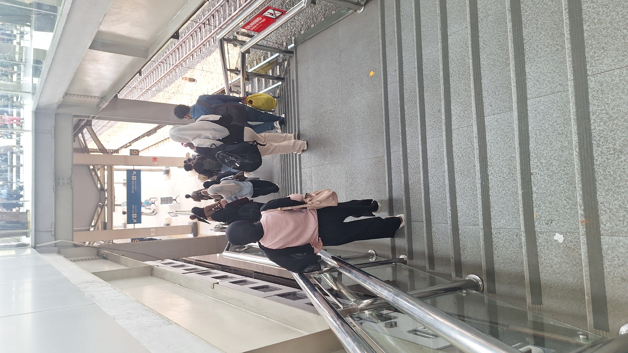 The image shows a group of people ascending a staircase at a train station in Cikarang, Indonesia. The stairs are gray and feature metal handrails with a glass panel. A sign indicating the train platform is visible overhead. The people are a mix of adults and young adults of varying ethnicities, wearing casual clothing, some with bags.