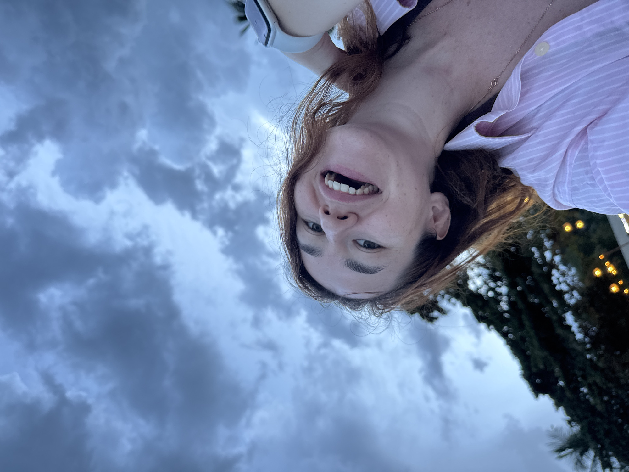 A close-up, upside-down selfie shows a woman with long, reddish-brown hair and freckles smiling broadly, revealing her teeth. She is wearing a pink and white striped button-down shirt and a gold necklace. A smartwatch is visible on her left wrist. The background is a mix of dramatic, cloudy skies and a blurred, dark green tree line with bokeh lights, suggesting an outdoor setting at dusk or during a cloudy day. The weather appears overcast. The overall emotion conveyed is cheerful and uninhibited. The location is identified as Baan Chaweng Noi, Thailand.