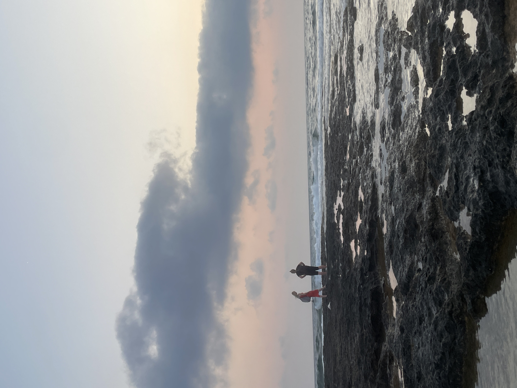 The image captures a serene coastal scene at sunset, likely along the rocky shores of Paphos Municipality, Cyprus. Two individuals, a woman in a red dress and a man in dark attire, stand on a dark, volcanic rock formation that extends into the sea. The woman is facing left, and the man is facing right with his hands on his hips, suggesting they are observing the ocean. Small pools of water are visible amongst the rocks, reflecting the soft hues of the twilight sky. The sea is moderately active with gentle waves rolling towards the shore. The sky above transitions from a pale blue at the top to a soft pink and orange as it nears the horizon, with scattered, dark clouds adding texture. A single, faint star is visible in the upper portion of the sky. The overall atmosphere is tranquil and contemplative, characteristic of a coastal sunset.