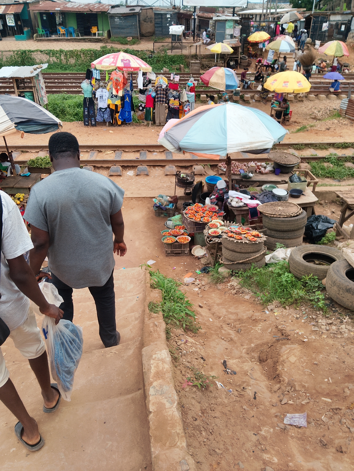 The image depicts an outdoor market scene, likely in Ijoko, Nigeria, situated next to railway tracks. The market appears to be a vibrant, bustling place with numerous stalls and vendors. In the foreground, two individuals are walking down a set of dirt steps, one carrying a clear plastic bag filled with a coiled blue rope. Behind them, a variety of market stalls are visible, displaying clothing, food items, and other goods.
The market stalls are set up under colorful umbrellas, providing shade for the vendors and their merchandise. One stall displays a selection of T-shirts and shorts. Another stall features an assortment of food items, including what appear to be tomatoes and other produce arranged in baskets and bowls. The ground is uneven and dusty, typical of an outdoor market environment.
In the background, more stalls and structures are visible, some of which appear to be makeshift shops. People can be seen browsing at the stalls, interacting with vendors, and moving about the market. The railway tracks are a prominent feature, suggesting the market's location near transportation infrastructure. The overall impression is one of a busy, local marketplace, indicative of daily life and commerce in the area.