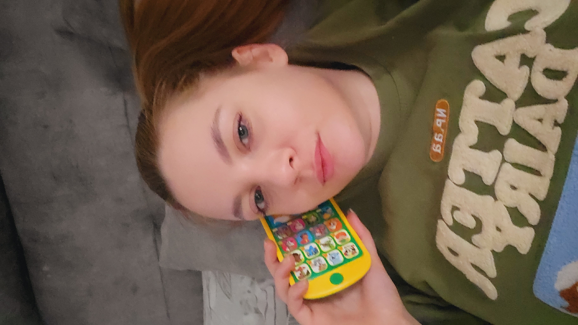 A woman lies on a gray upholstered surface, possibly a sofa or bed. She has fair skin, reddish-blonde hair pulled back, and is wearing a green sweatshirt with cream-colored lettering and a brown rectangular patch with numbers. She is holding a yellow toy phone with colorful buttons, positioning it near her right eye. The photograph appears to be taken indoors. There is no visible information in the image to confirm the location in the city of Chaikovsky, Russia.