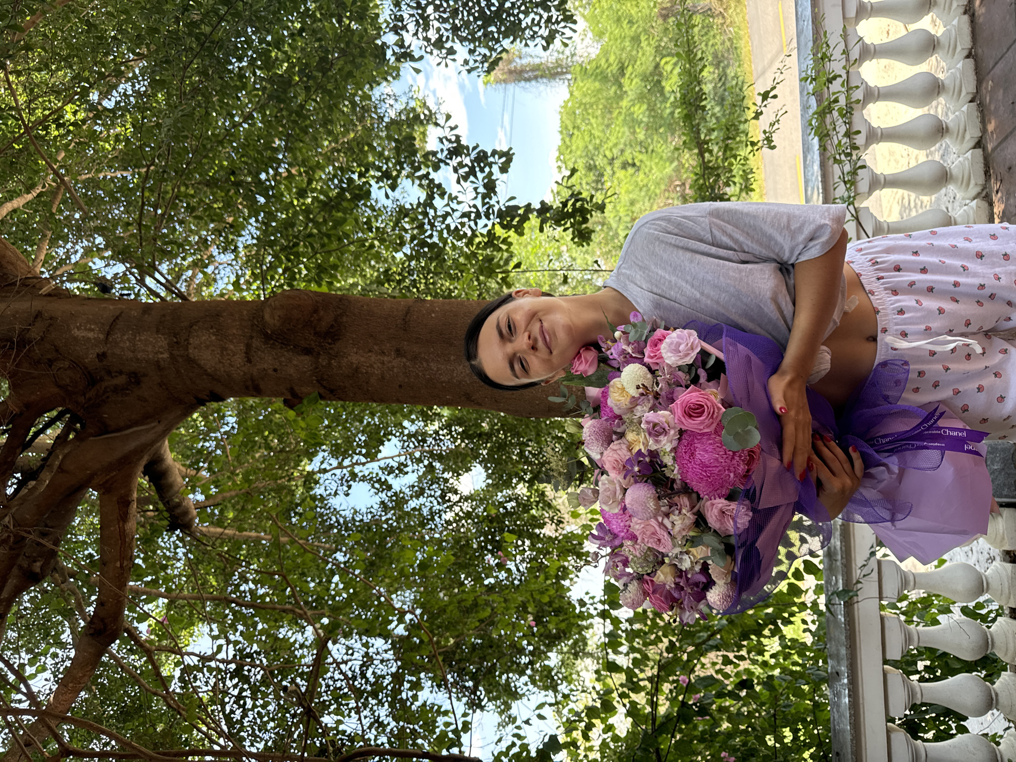 A young woman stands outdoors, holding a large bouquet of pink and purple flowers. She is wearing a grey t-shirt and patterned shorts. Behind her, a large tree with green foliage and a blue sky are visible. The scene appears to be on a balcony or porch, with a white railing in the foreground. The location is identified as Mũi Né, Vietnam, suggesting a tropical or subtropical environment.