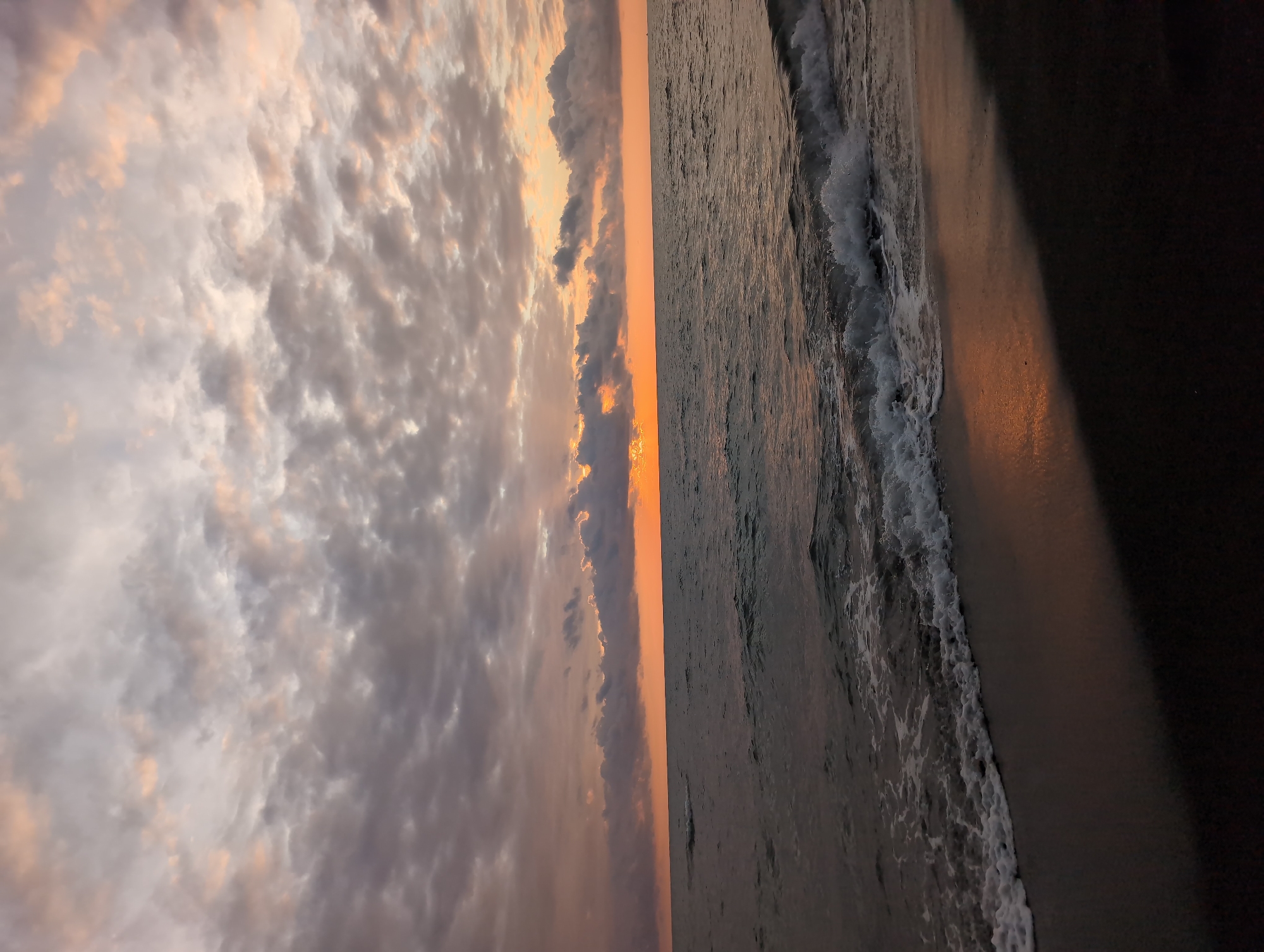 This image captures a twilight scene at the beach in Ureki, Georgia. The sky is filled with a mix of gray and pinkish-orange clouds, indicating either sunrise or sunset. The sea is dark and moderately choppy, with small waves breaking on the shore. The beach itself is dark, possibly wet sand or pebbles. The overall impression is one of a calm, natural environment at the end or beginning of the day. There are no people or man-made objects visible in the frame.