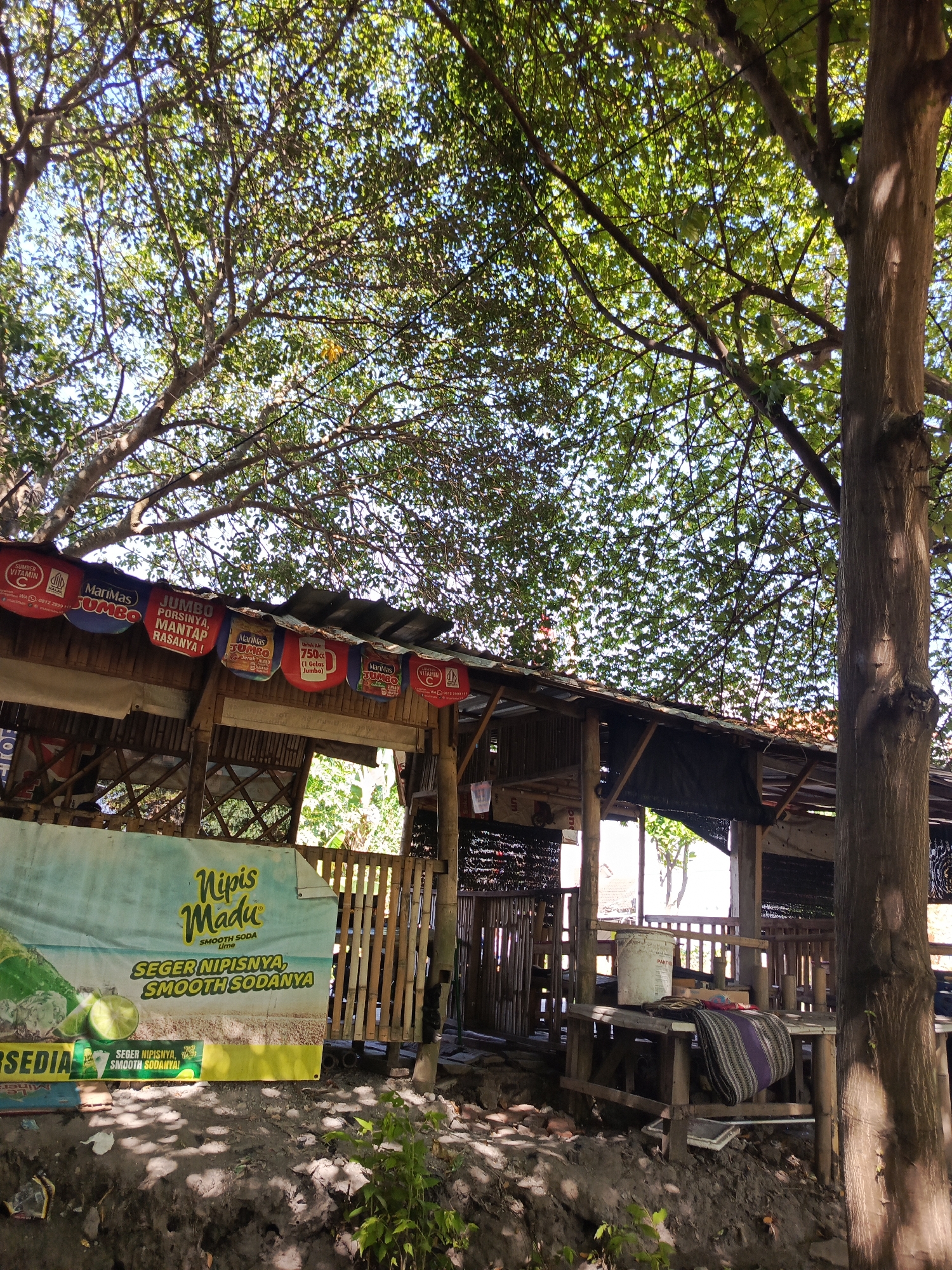 This image captures a rustic roadside stall or eatery under the shade of large trees, likely located in a rural or semi-urban area of Kediri City, Indonesia. The structure is primarily made of wood and bamboo, with a corrugated metal roof. Several colorful banners advertising beverages, including "Nipis Madu" (which appears to be a lime-flavored soda) and various "Jumbo" drinks, are displayed along the front of the stall. A large banner with an image of limes and text in Indonesian promotes the "Nipis Madu" product, stating "SEGER NIPISNYA, SMOOTH SODANYA" (Its freshness is limey, its soda is smooth). In the foreground, the ground is uneven and appears to be dirt with some scattered debris. A small plant is growing in the lower right corner. A wooden table with a white bucket and a striped cloth is visible within the stall. The overall impression is of a simple, local establishment operating in a natural, sun-dappled environment. There are no people visible in the image.