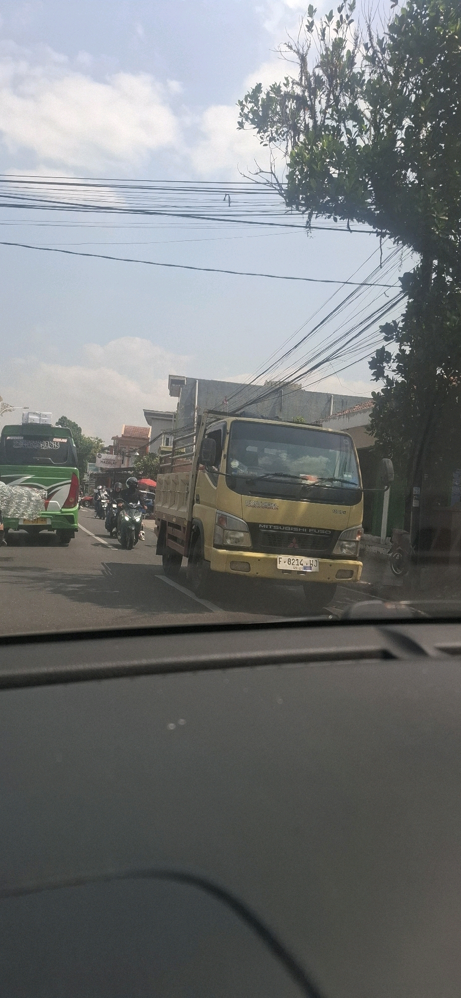 The image shows a street scene in Sukasari, Indonesia, on a sunny day with a partly cloudy sky. In the foreground, the interior of a vehicle is visible, with a dark dashboard and reflections on the windshield. The main focus of the image is a yellow Mitsubishi Fuso truck with a flatbed, driving in traffic. The truck's license plate is visible and reads "F 8214 HJ". To the left of the truck, a green bus is also in traffic. Several motorcycles are also present on the road, with riders. Buildings line the side of the street, some appearing to be shops or residences. Overhead, a network of power lines is strung across the scene, with trees and branches visible on the right side of the image. The road is paved and appears to be a busy thoroughfare.