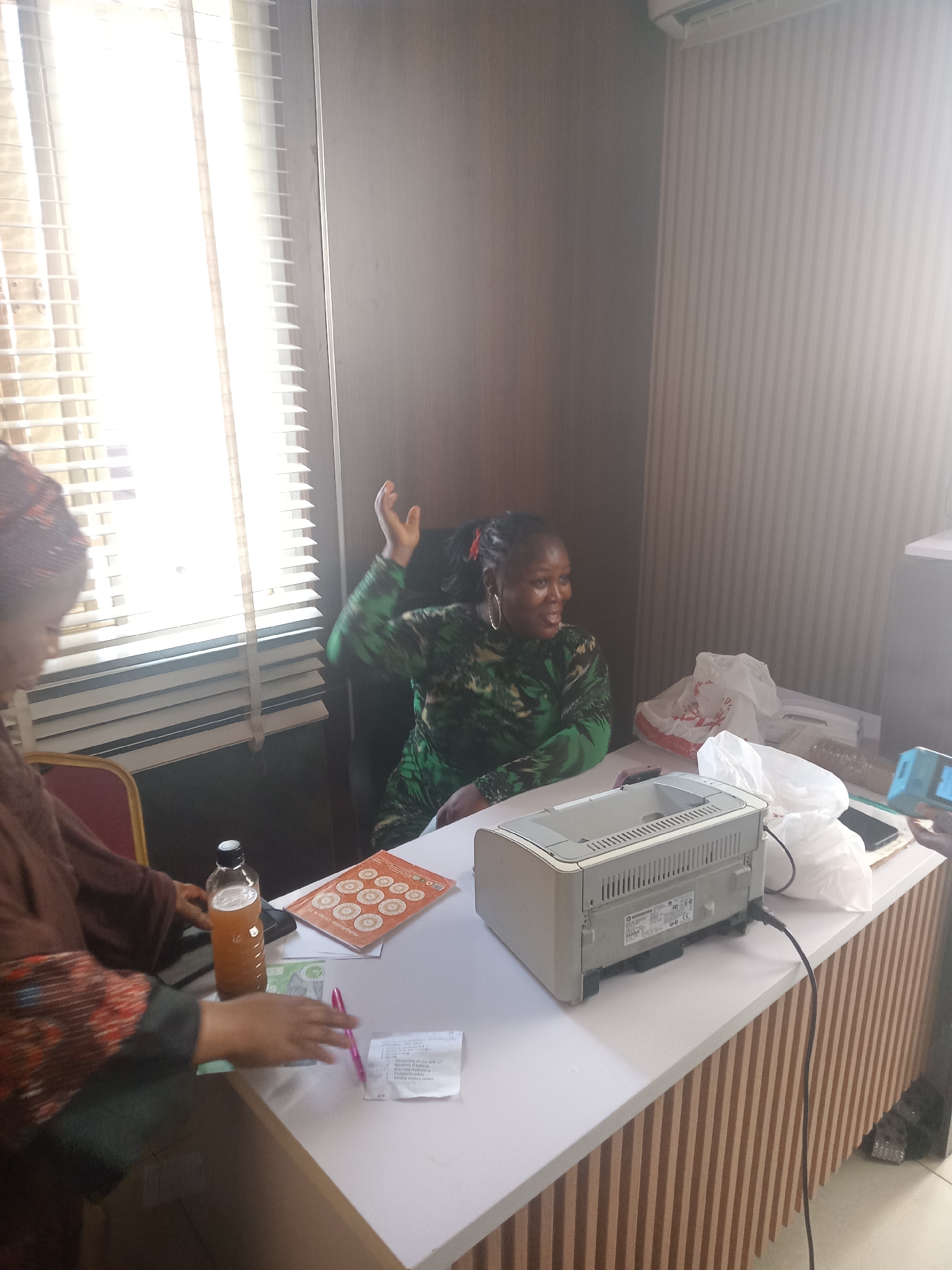 Two women are in a room in Gwarinpa, Nigeria. One is partially visible on the left, wearing a patterned brown top and head covering, holding a pink pen near a receipt, a bottle of liquid, and a brochure. The other woman sits in a chair, wearing a green patterned top, with her right hand raised. On the desk in front of them is a printer, a plastic bag, a notebook, and a mobile phone. The room has a window with blinds on the left and light-colored striped wallpaper.