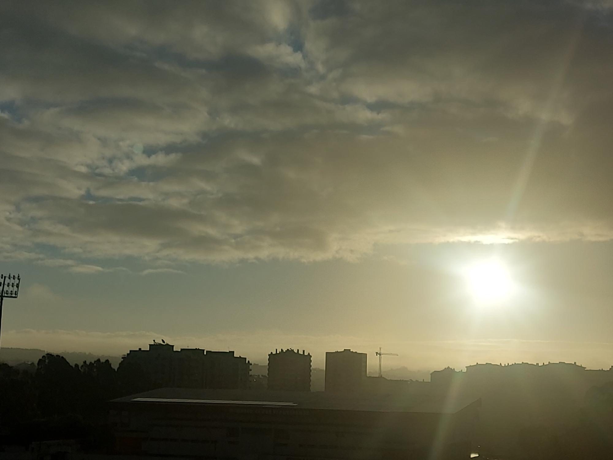 The image shows a cityscape at dawn or dusk in Figueira da Foz, Portugal. The sun is bright and low in the sky, shining through a layer of clouds. In the foreground, there are several buildings, including residential blocks and what appears to be a stadium or large structure. A crane is visible amidst the buildings in the middle distance. A tall light pole is located on the left side of the image. The overall atmosphere is hazy due to the bright sunlight and atmospheric conditions.